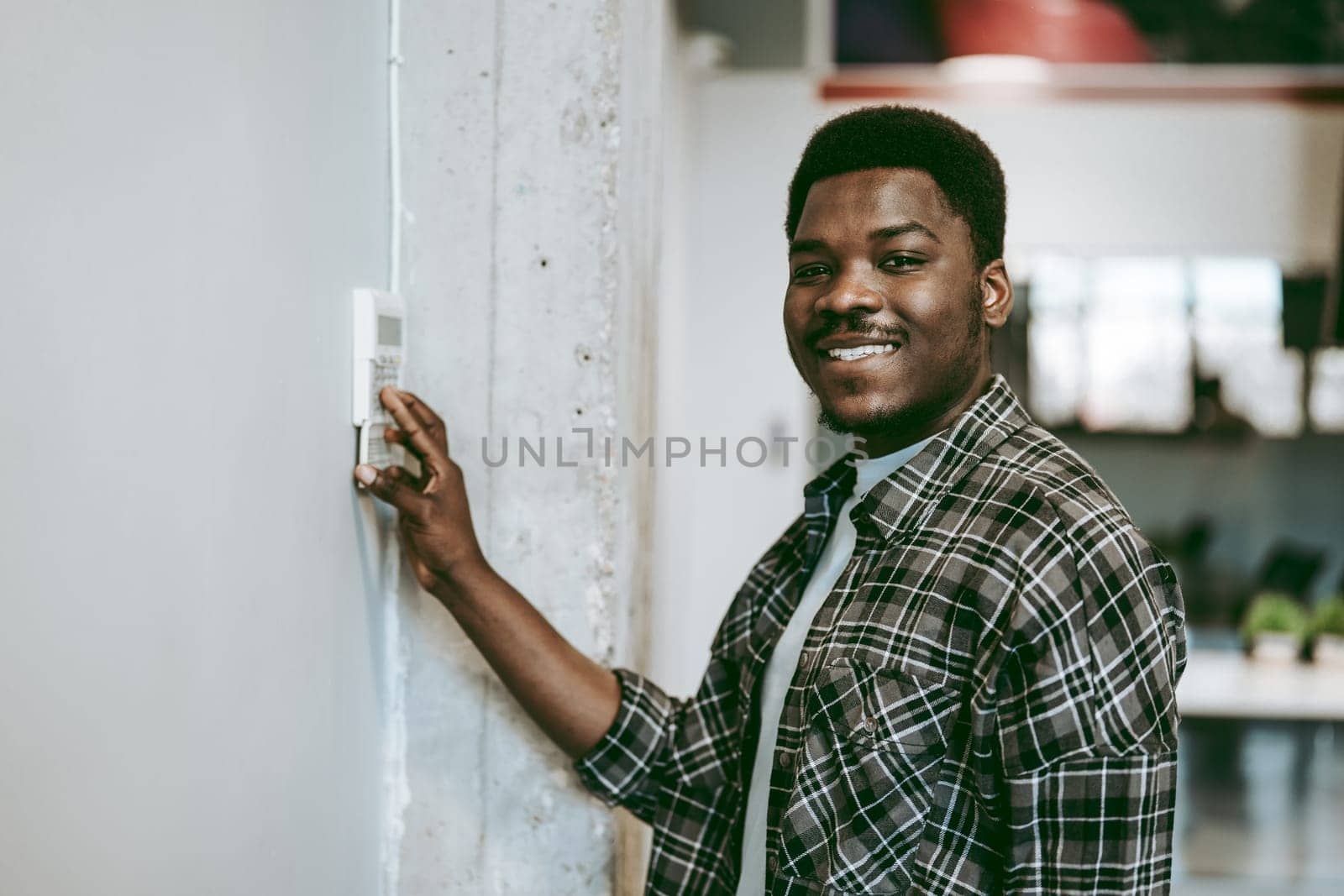 A young man with a cheerful expression is standing in a contemporary office, adjusting the thermostat on the wall. Sunlight fills the space, enhancing the positive atmosphere.