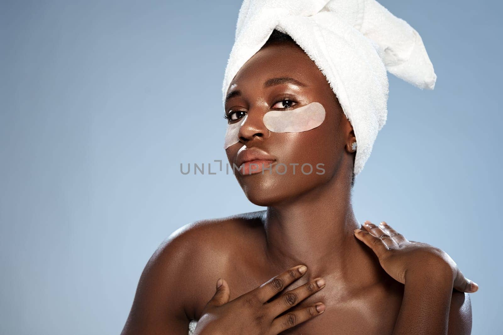 A woman with dark skin and a towel on her head applies a soothing face mask while standing against a light blue background. She embodies relaxation and self-care.