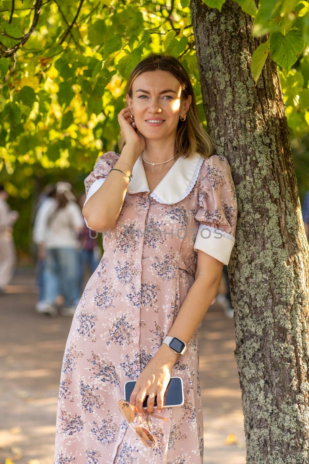 A woman stands gracefully by a tree at Peterhof, dressed in a floral dress, capturing the essence of a bright and lively outdoor day with visitors nearby.