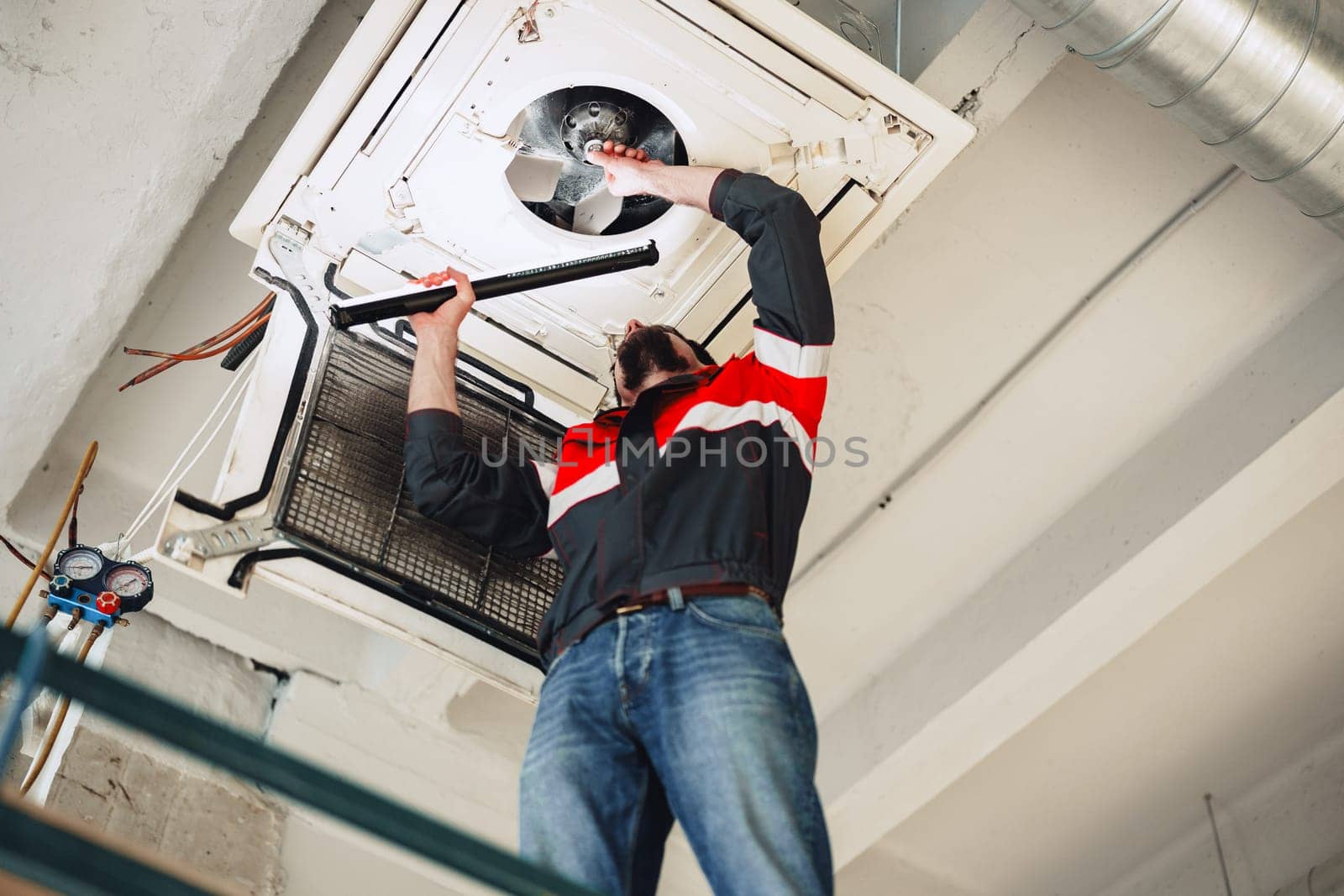 A technician is inspecting and repairing an air conditioning unit on the ceiling of a commercial building in the afternoon. Tools are visible, and the technician shows focus and concentration.
