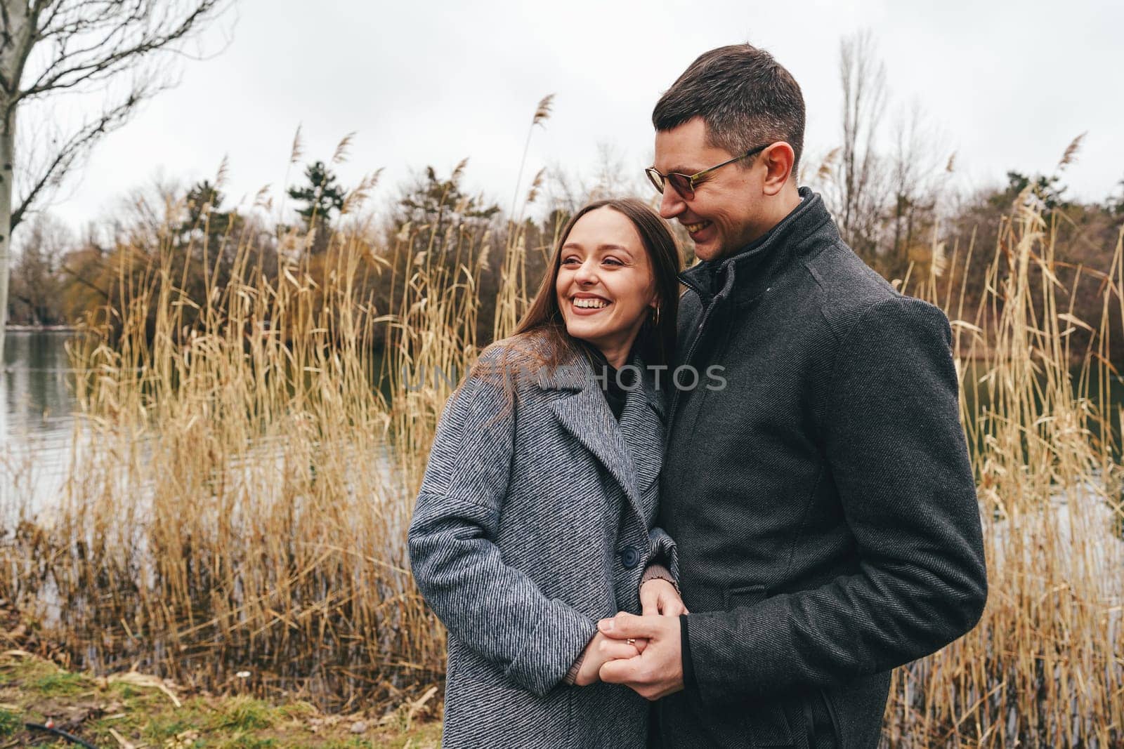 Two people share a joyful embrace near a tranquil lake, surrounded by tall grasses on a cool autumn day. Their smiles reflect happiness and warmth amidst the overcast sky.