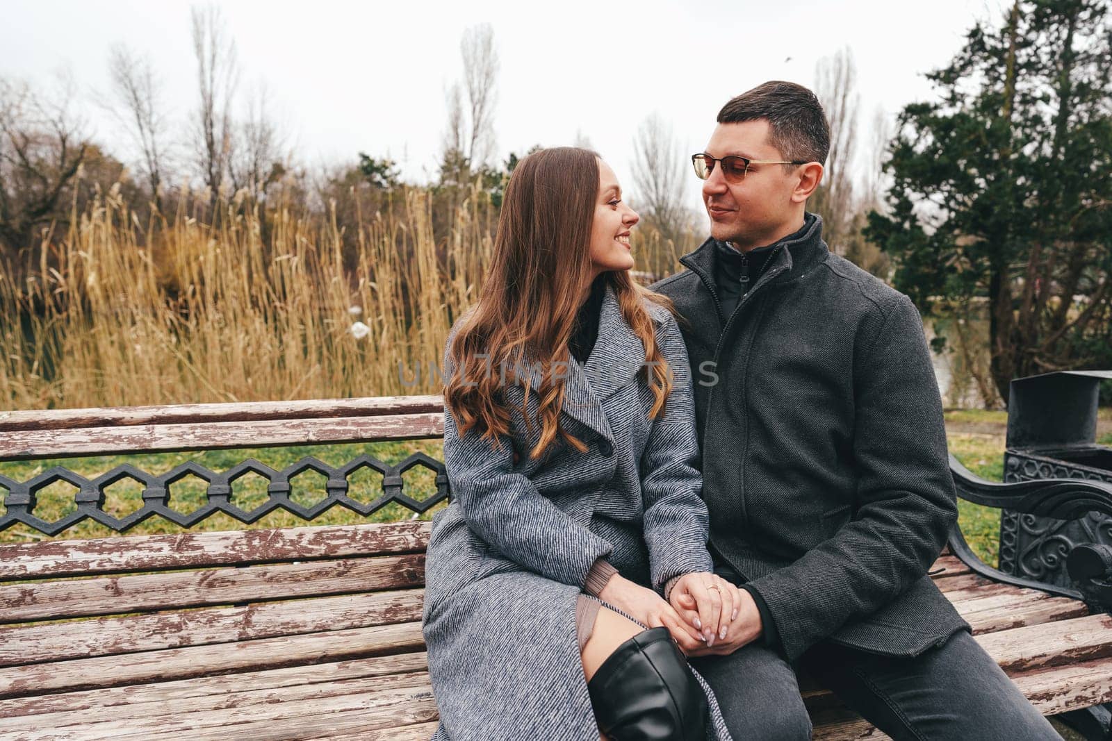 A couple sits closely on a bench in a park, sharing smiles as they enjoy a chilly afternoon in late fall surrounded by tall grass and trees.