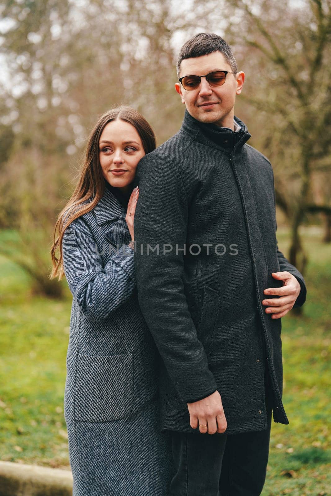 A couple stands closely together in a peaceful park, dressed warmly for the cold weather. The woman rests her head affectionately on the man's back, surrounded by trees.