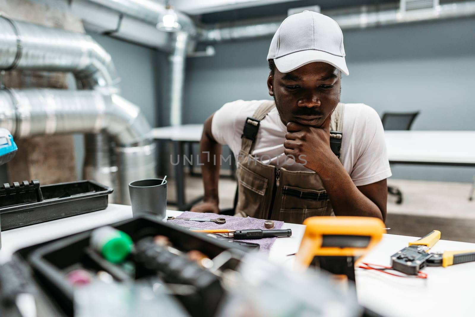 A young man is focused on a collection of tools and equipment laid out on a table in a workshop. He appears to be thinking critically about the task at hand.