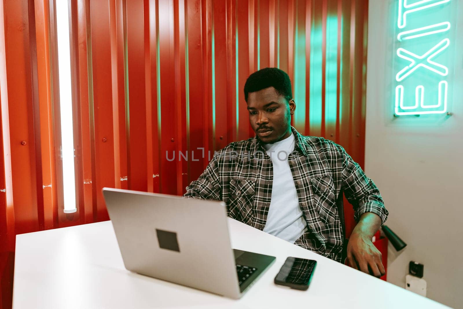 A young man is focused on his laptop in a trendy coworking space characterized by bright red walls and a glowing green exit sign. He appears engaged in his work.