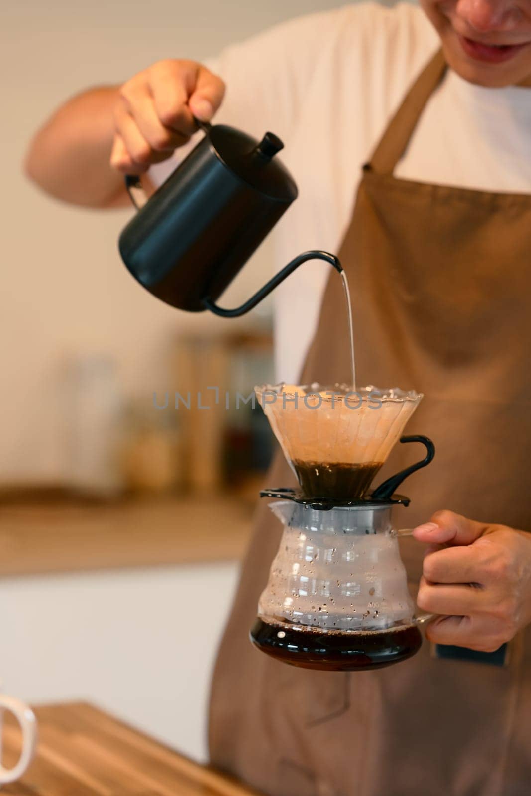 Young man in apron brewing coffee using the pour-over method in warm kitchen.
