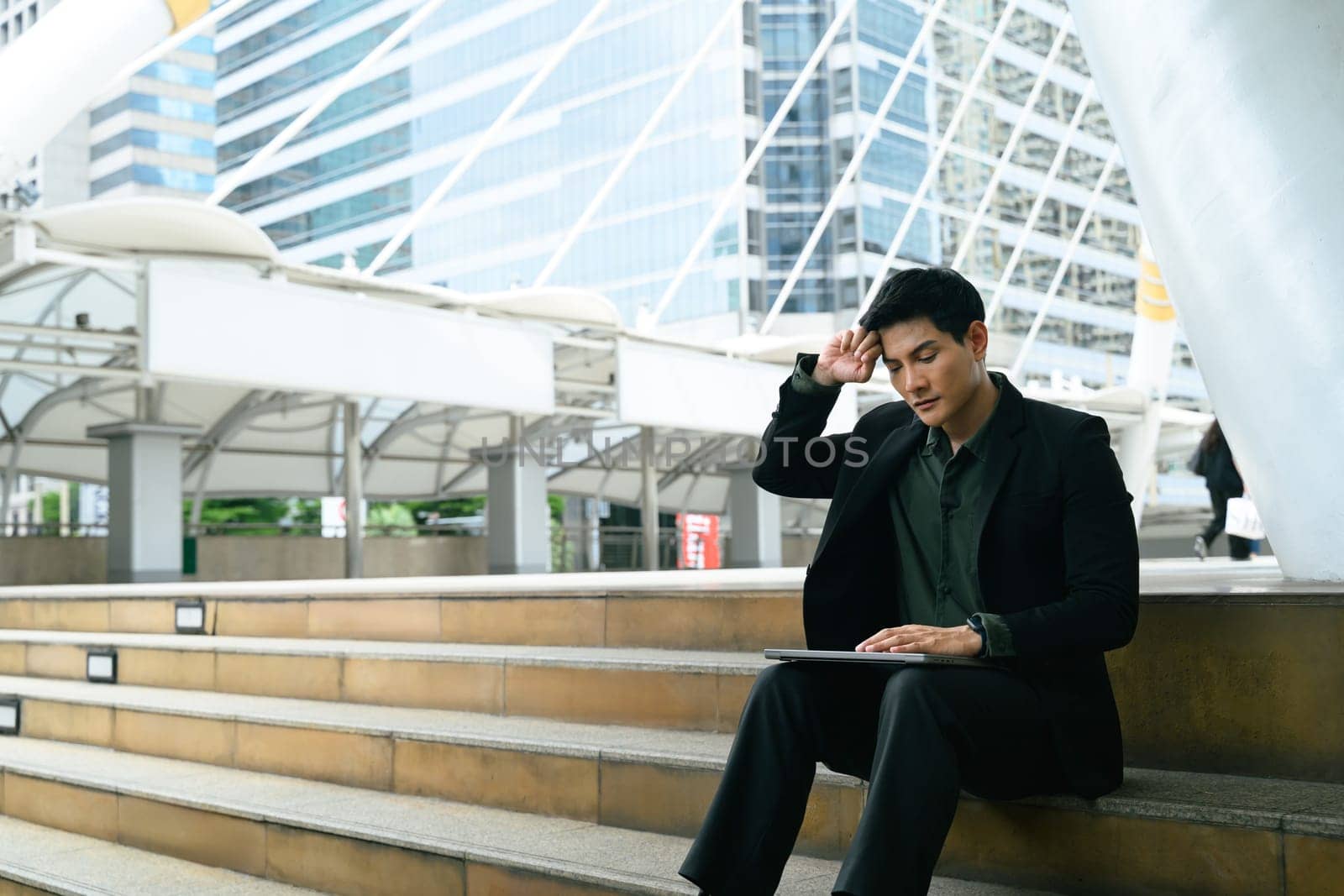 Exhausted businessman sitting on steps in a city, working remotely on a laptop..
