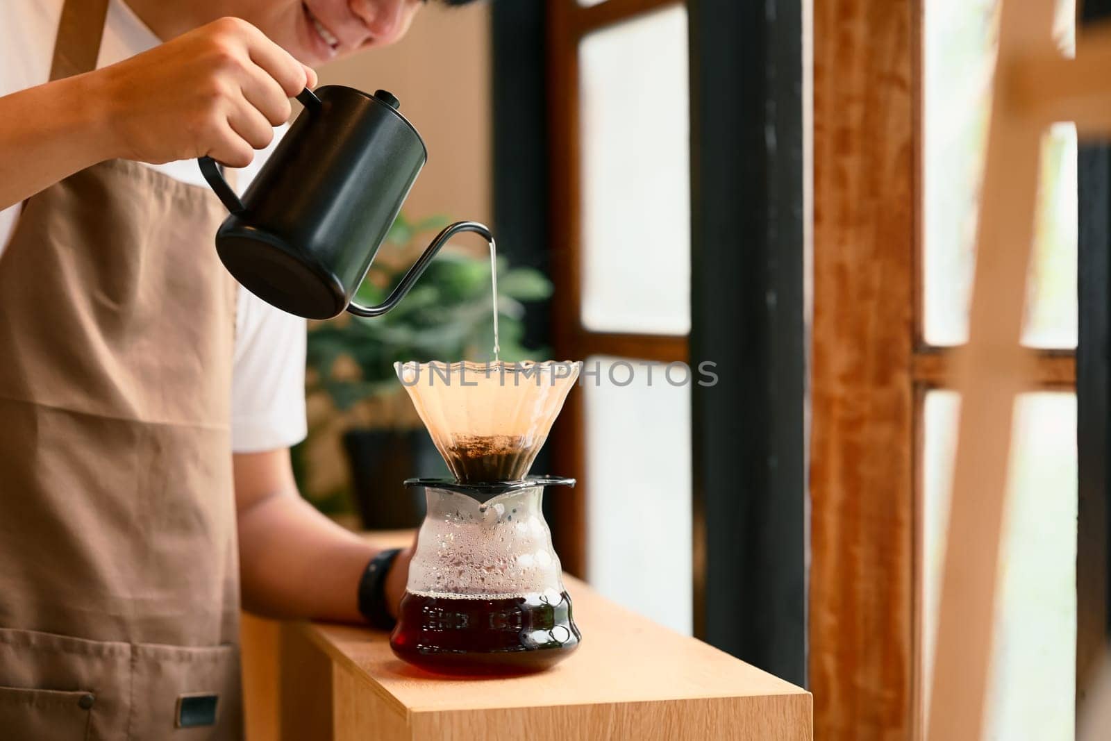 Young man in apron brewing coffee using the pour-over method in warm kitchen.