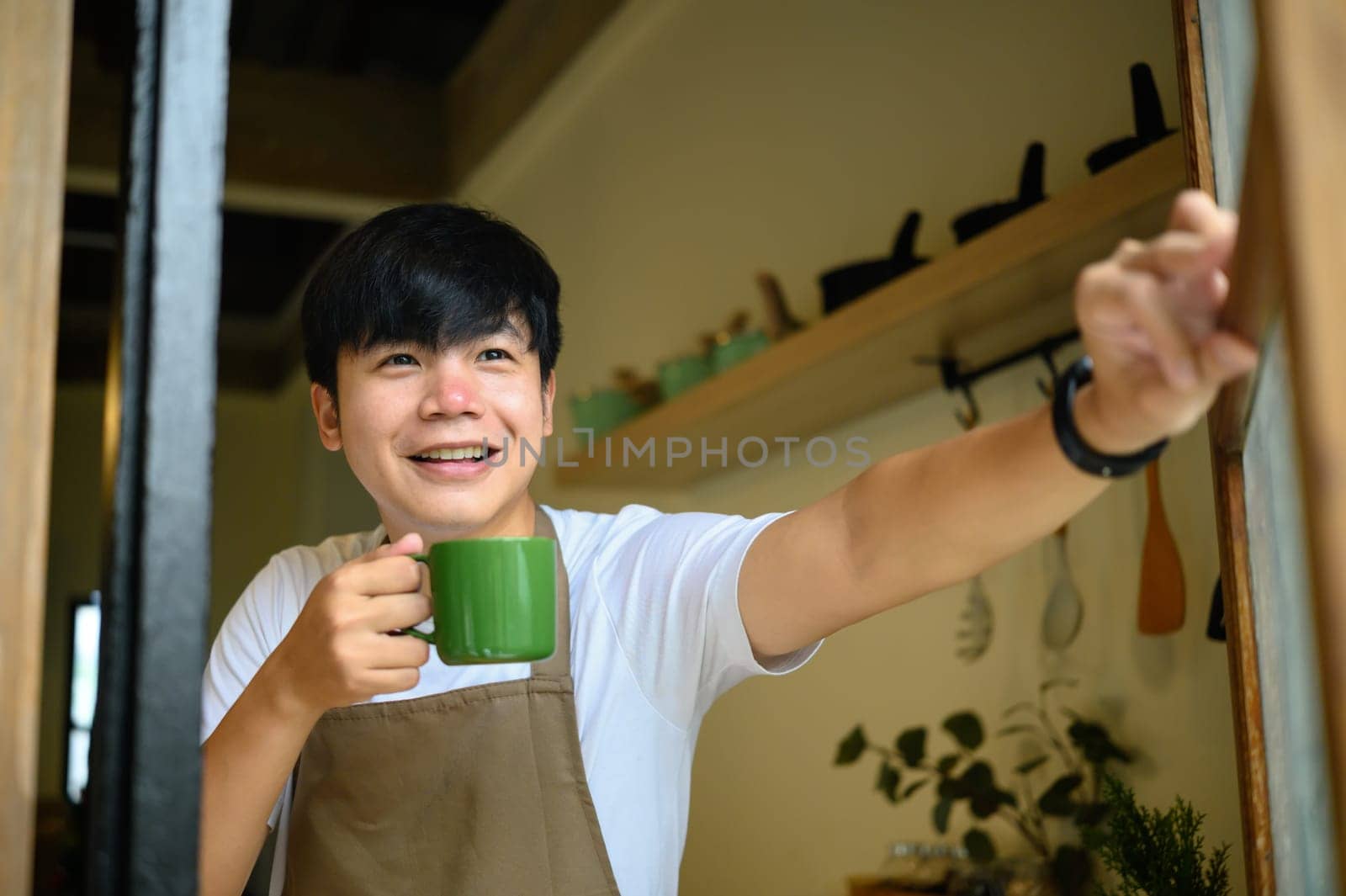 Young man holding a coffee cup smiling warmly as he opens a window in a cozy kitchen.