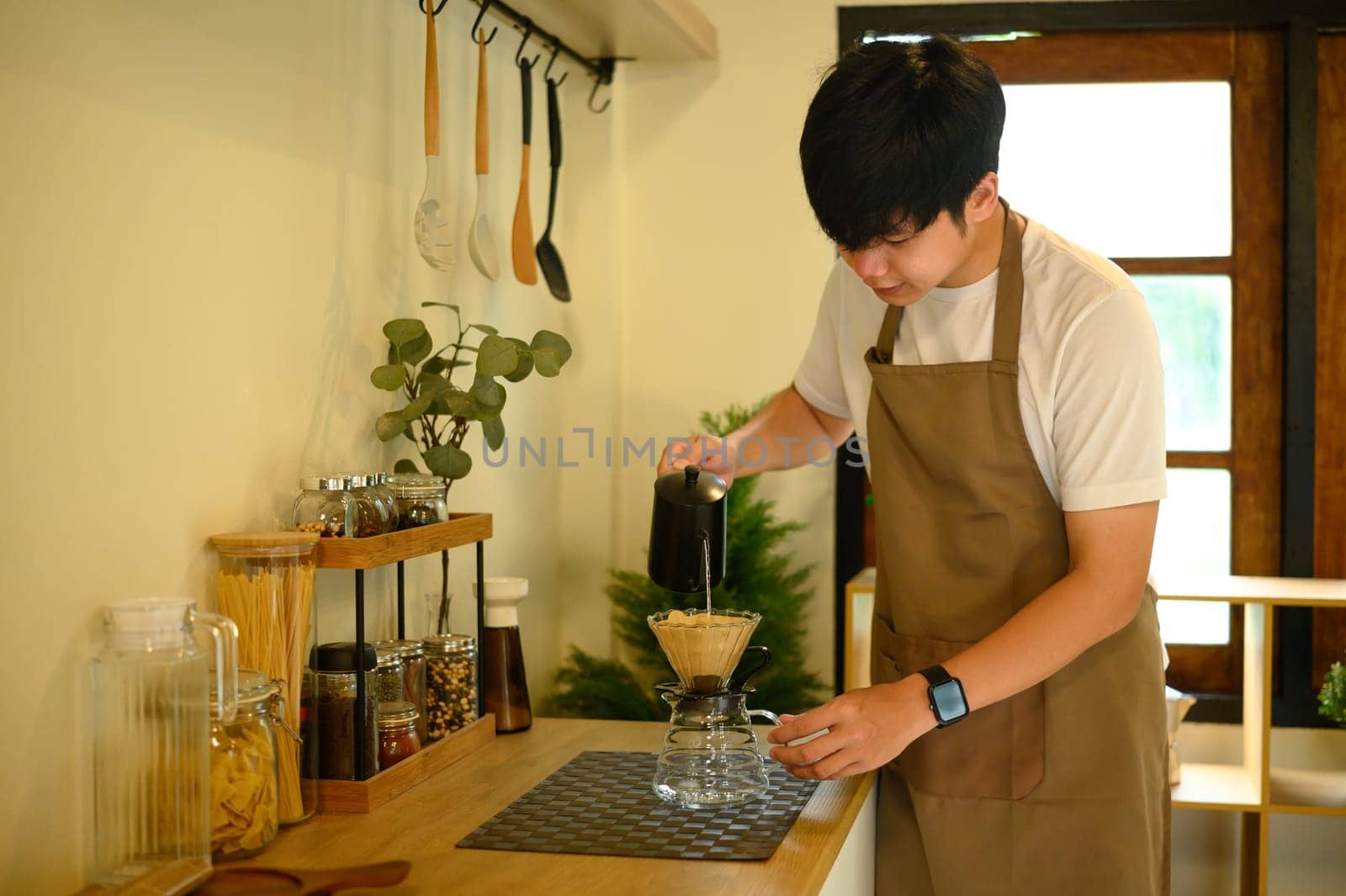 Asian man preparing a fresh cup of drip coffee in a modern, organized kitchen.
