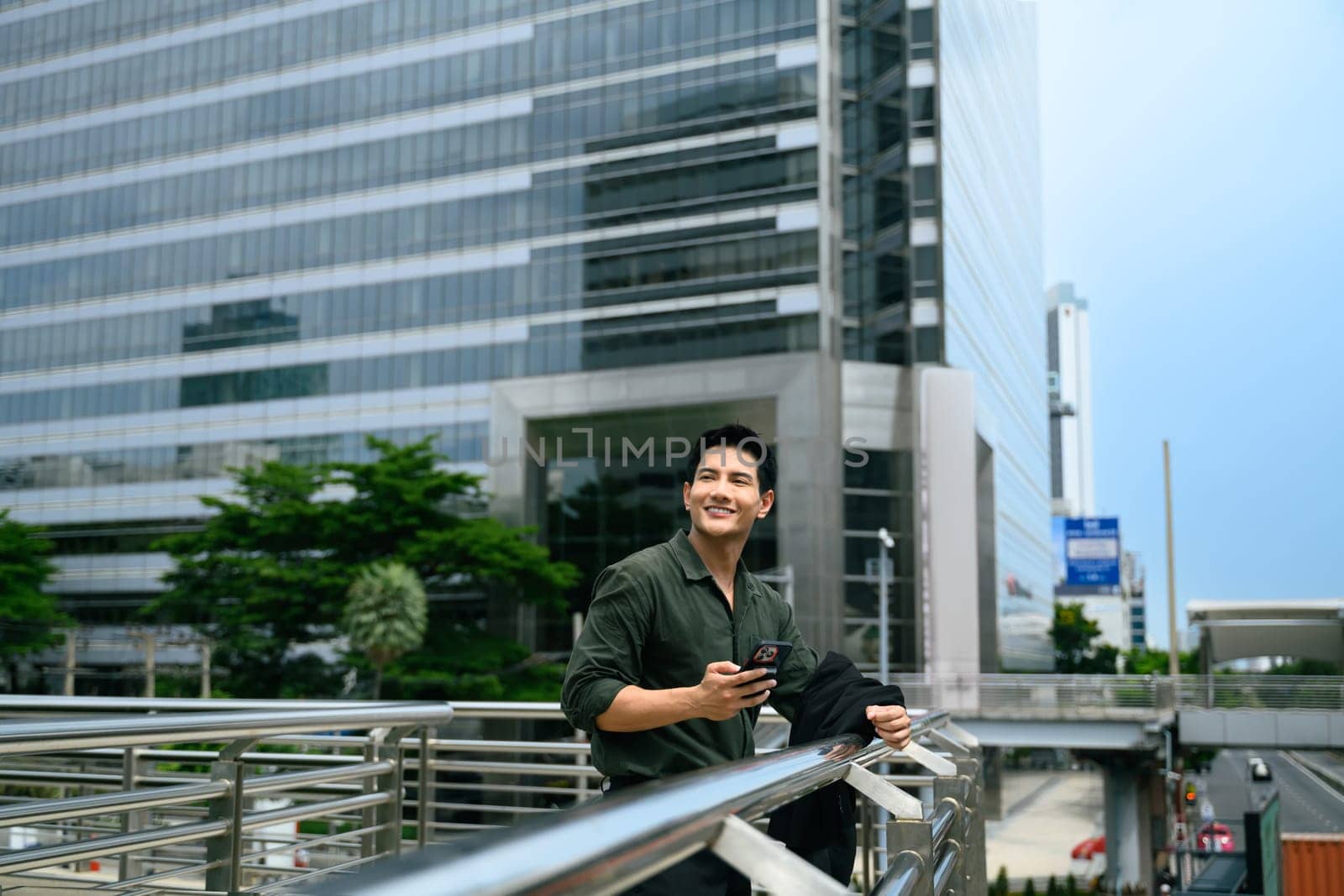 Smiling businessman holding phone standing by railing in a business district.