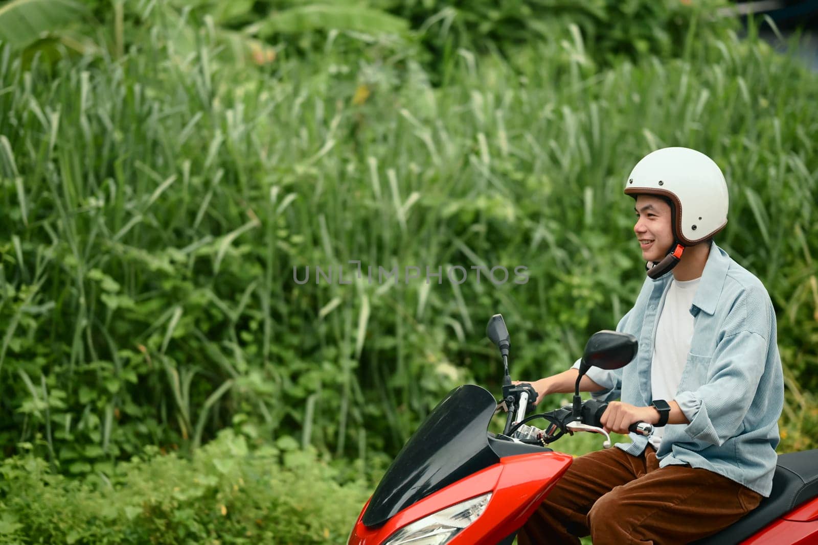Smiling young man in helmet riding a red motorbike along a rural road.