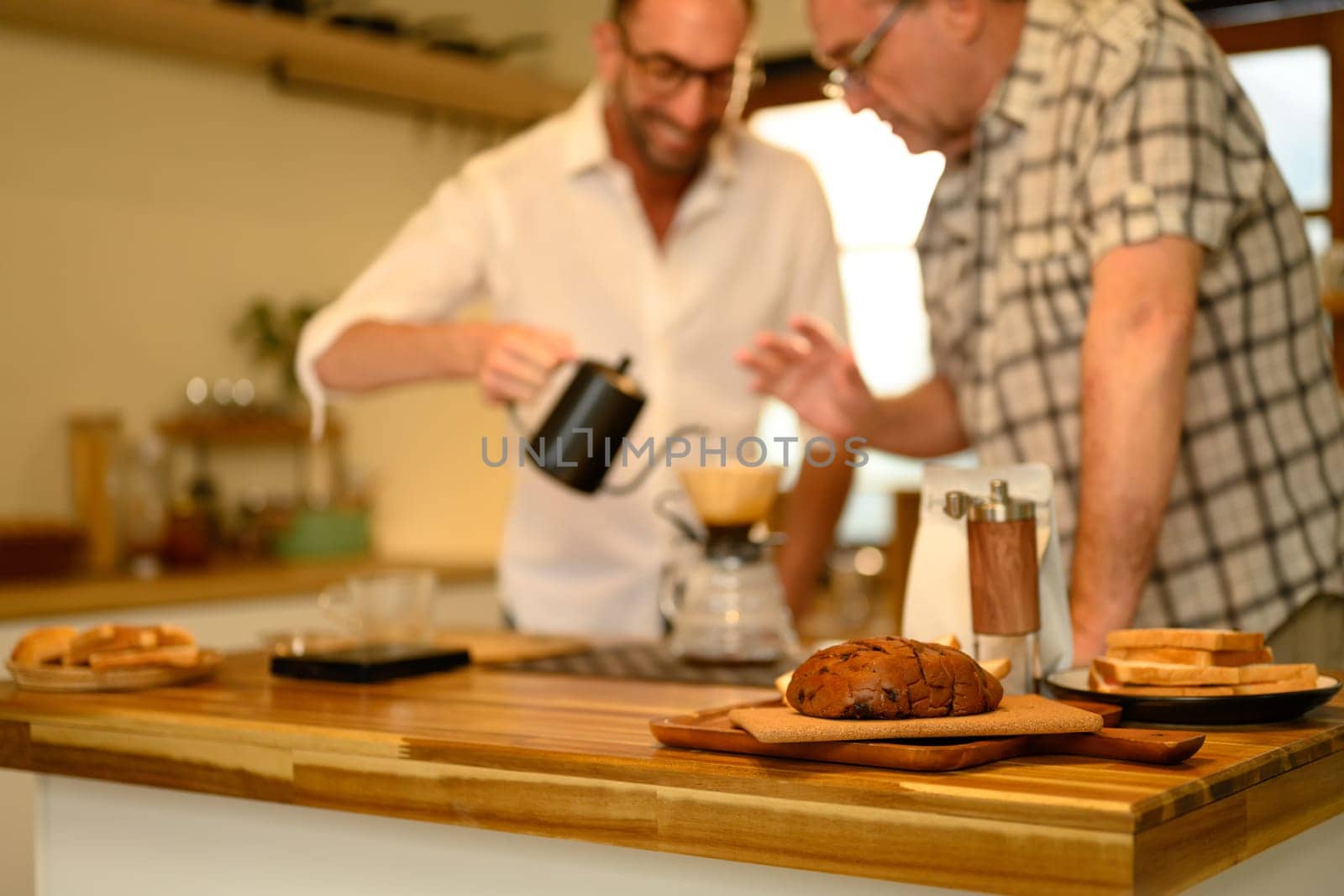 Sliced loaf of bread on a wooden countertop with father and son preparing coffee in background.