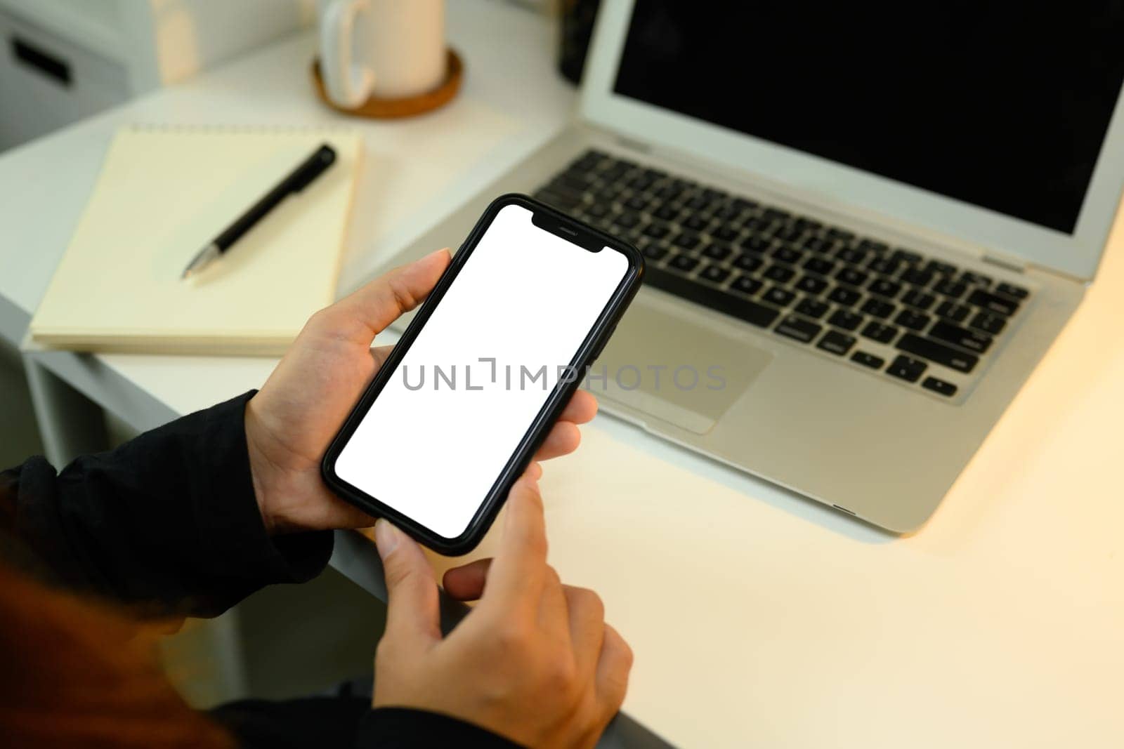 Close up of hands holding smartphone with blank white screen next to laptop.