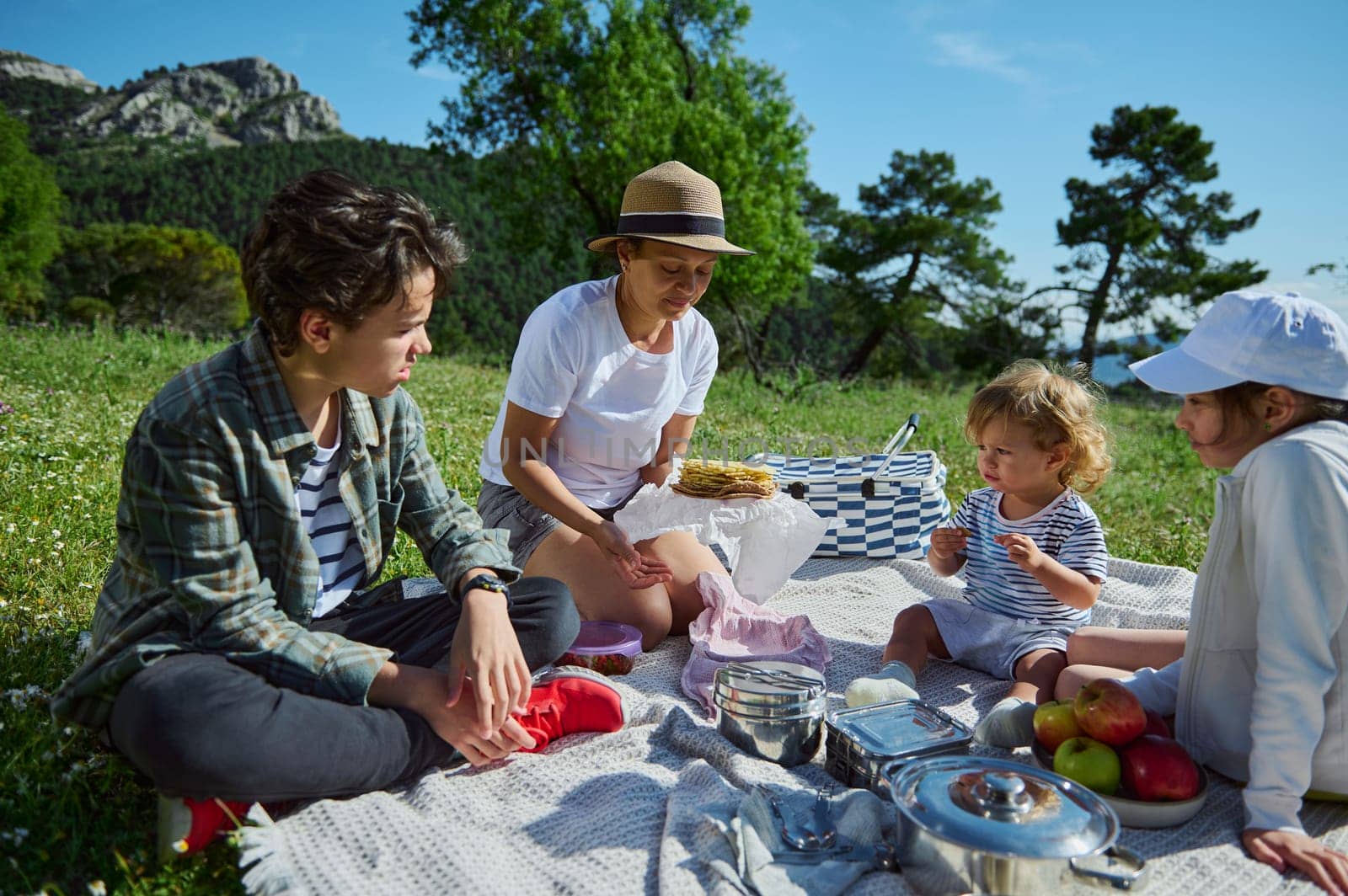 A warm family outing as a mother and her children gather for a picnic amidst beautiful mountain scenery and vibrant greenery under the summer sun.