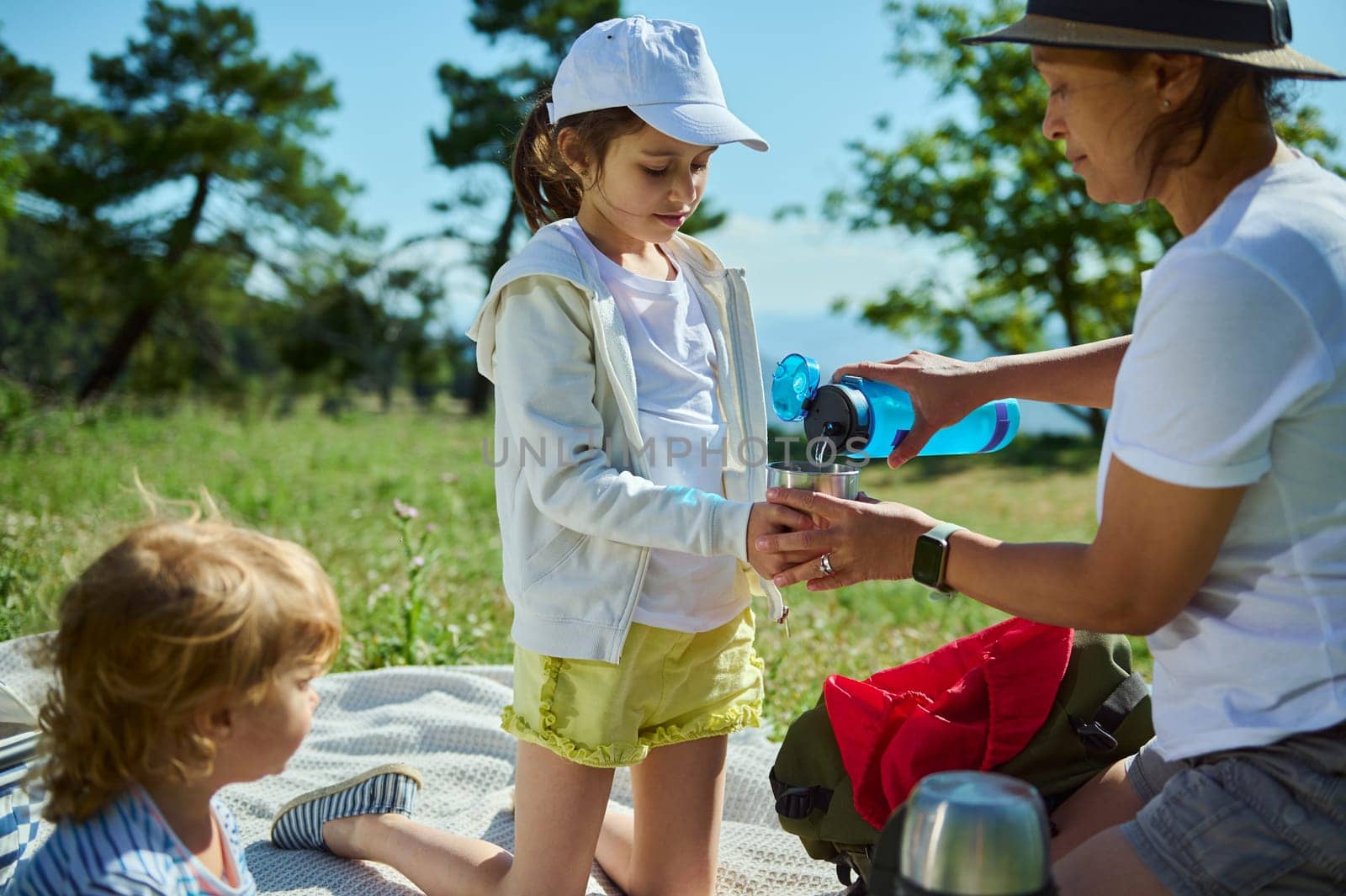 A mother pours a drink for her children during a sunny picnic in the mountains, emphasizing family bonding, outdoor leisure, and the beauty of nature.