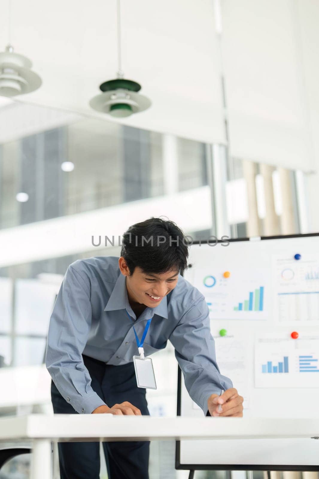 A young businessman smiles while taking notes on a whiteboard, reviewing business data in a modern office.