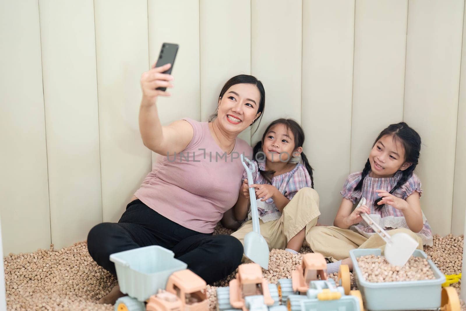 A joyful mother takes a selfie with her two daughters while playing with toys in a cozy playroom, highlighting family connection and fun.