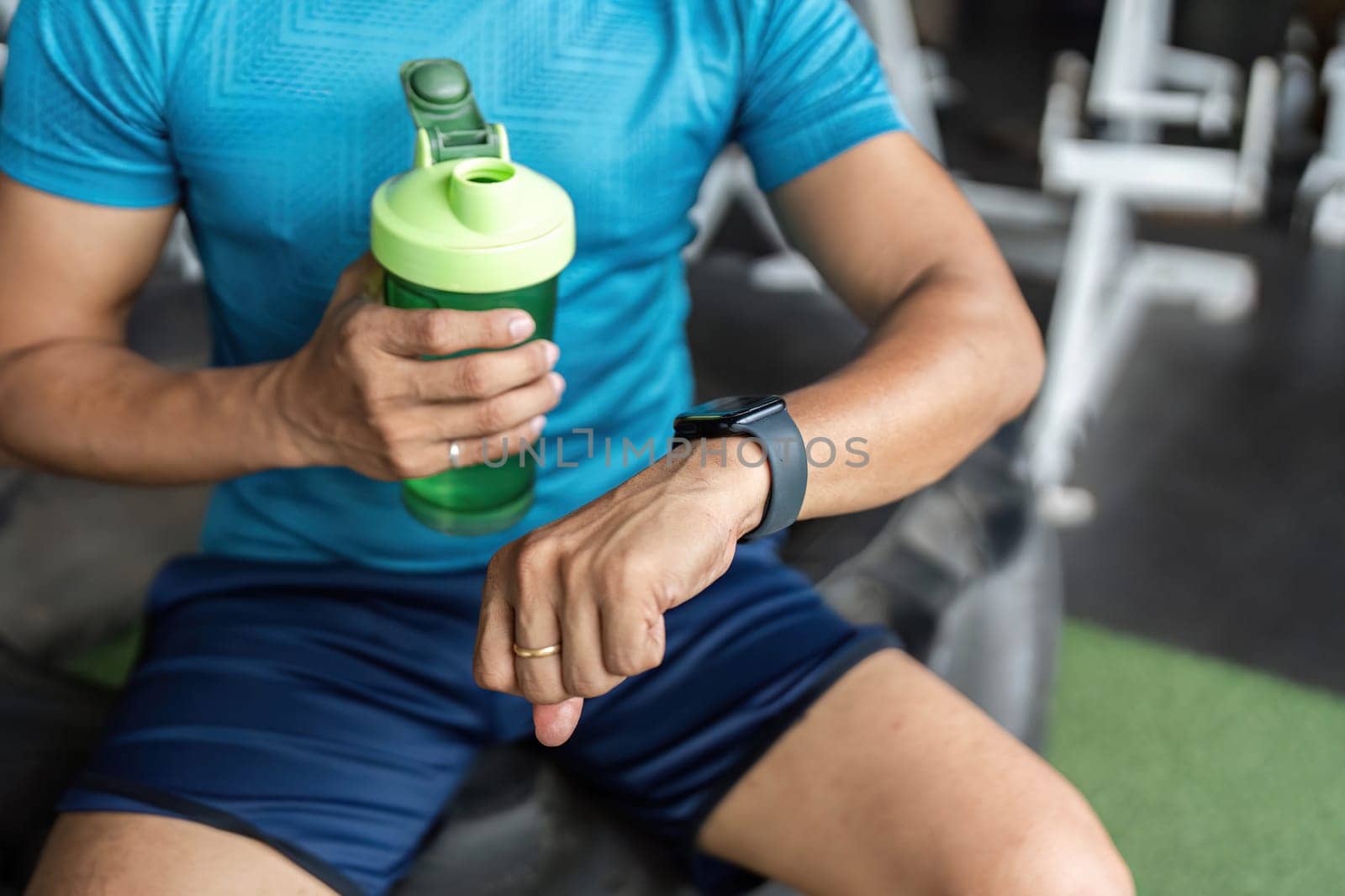 A fit man holding a green water bottle while checking his smartwatch during a workout session at the gym. Focus on fitness and hydration.