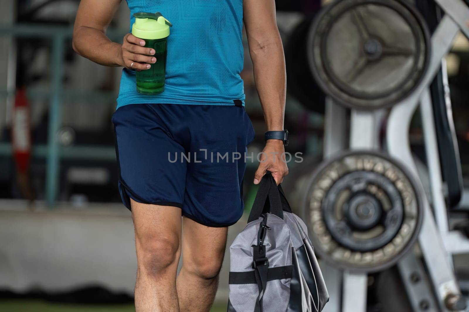 A man walking in a gym, carrying a shaker bottle and gym bag, showcasing a commitment to a fitness lifestyle. The mood is determined and focused.