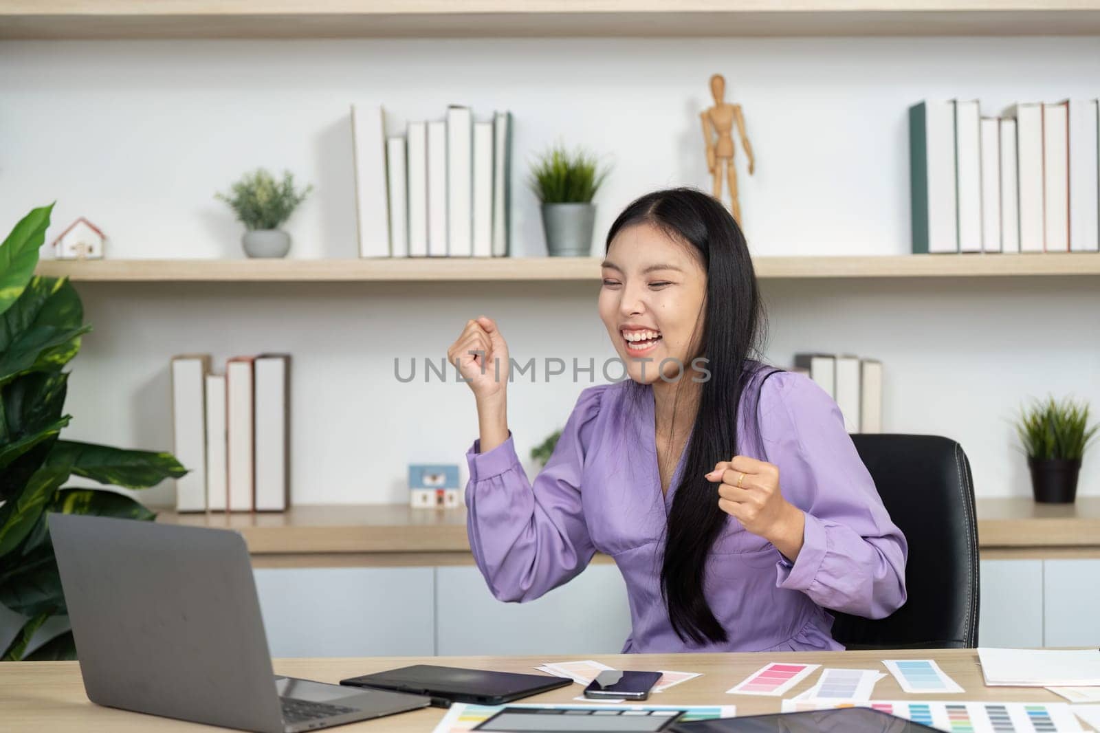 A joyful graphic designer celebrates a successful project at her workspace, surrounded by design materials and a laptop, showcasing enthusiasm and creativity.