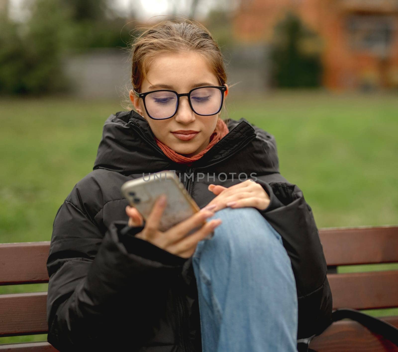 In Spring, A Girl In Glasses Sits On A Bench With A Smartphone