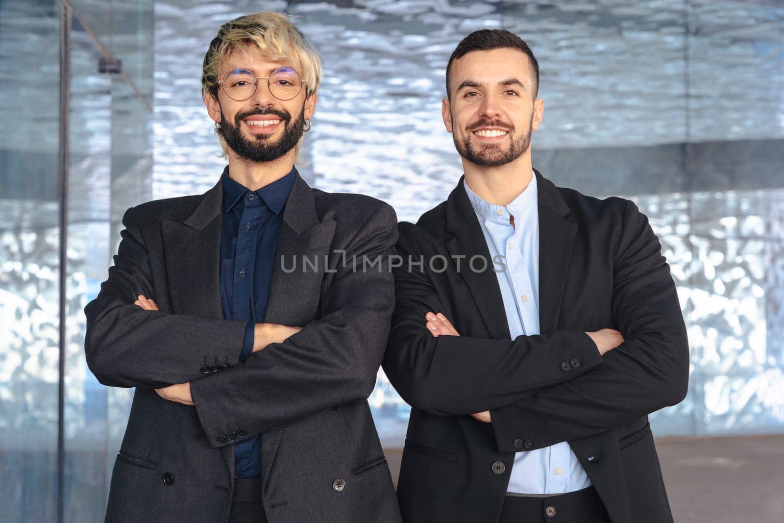 Two confident male professionals in suits standing with crossed arms, smiling at the camera inside a modern office. Corporate success, teamwork, and leadership in business environment.