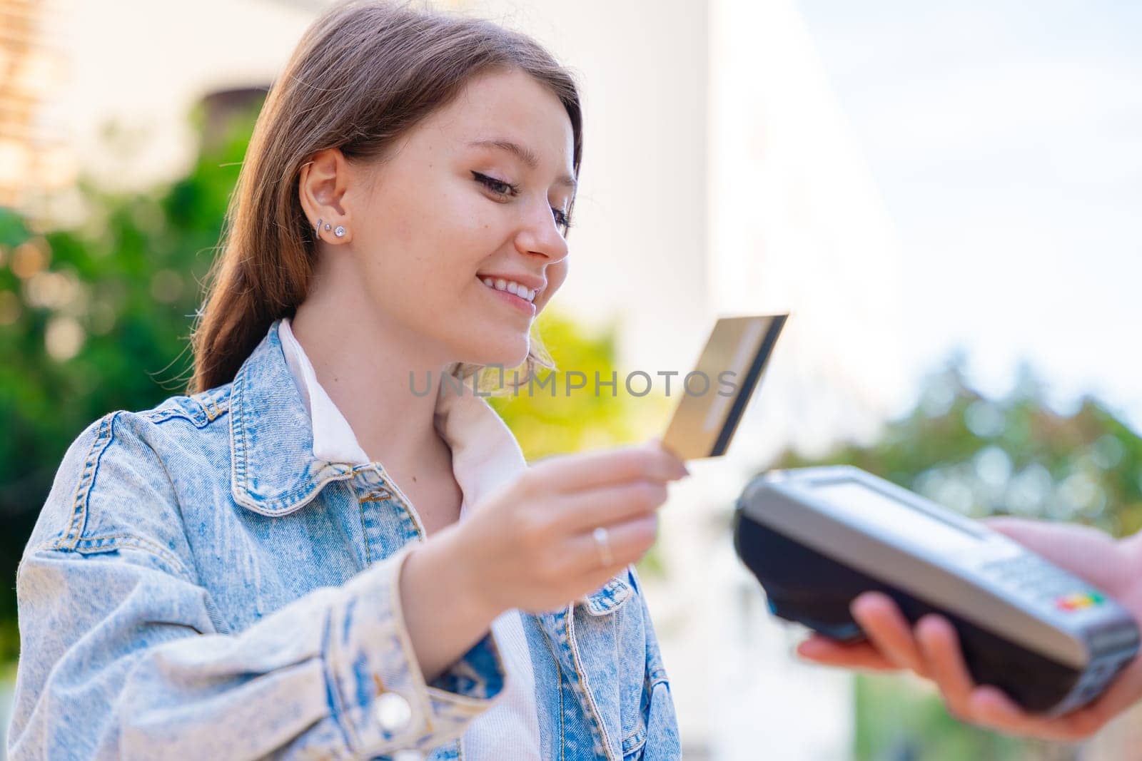 Caucasian woman smiling as she pays with a credit card using contactless technology at an outdoor cafe. A casual urban moment reflecting convenience, lifestyle, and modern financial habits.