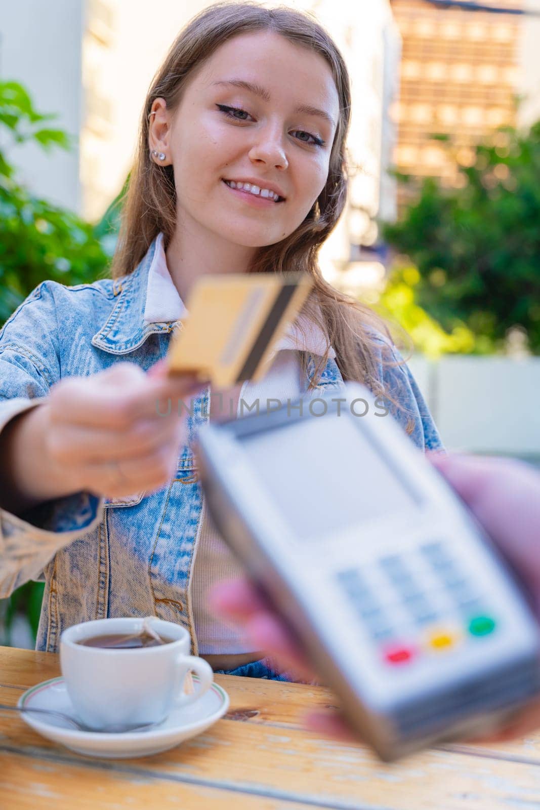Caucasian woman smiling as she pays with a credit card using contactless technology at an outdoor cafe. A casual urban moment reflecting convenience, lifestyle, and modern financial habits. Vertical