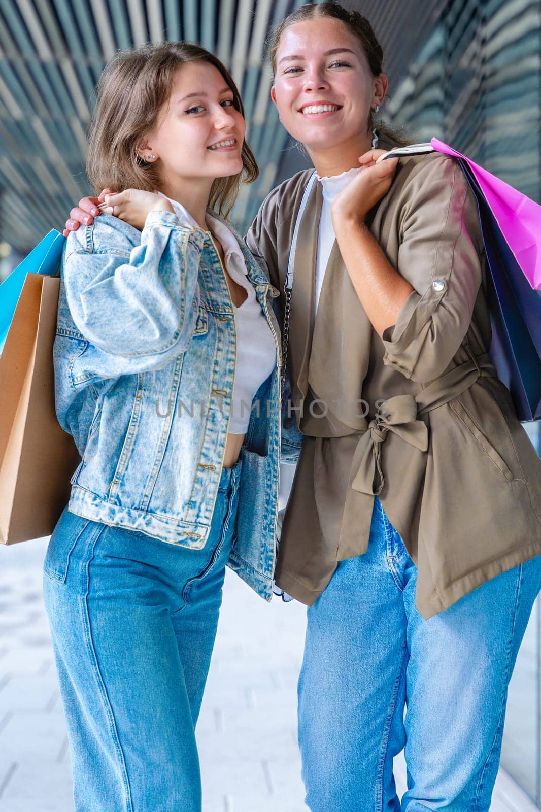 Two happy caucasian women holding colorful shopping bags and smiling after a successful day at the mall. They stand outside a modern building, enjoying their time together with style and joy. Vertical