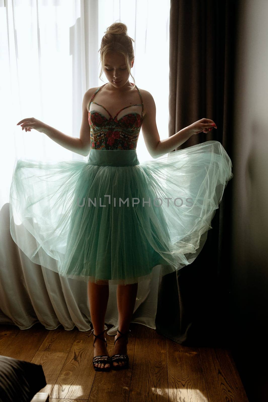 young blonde in turquoise dress stands near window in dress with full tulle skirt. Her skirt is transparent and flies. slender girl opposite window.