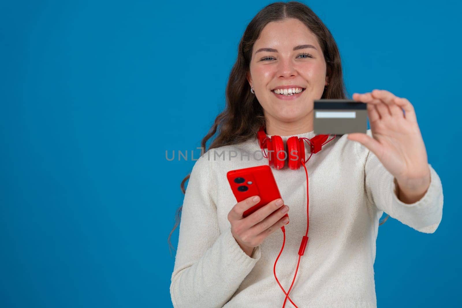 Happy young woman in a white sweater and red headphones smiling while showing a credit card to the camera and holding a red smartphone, against a blue background, symbolizing online shopping. Copy space