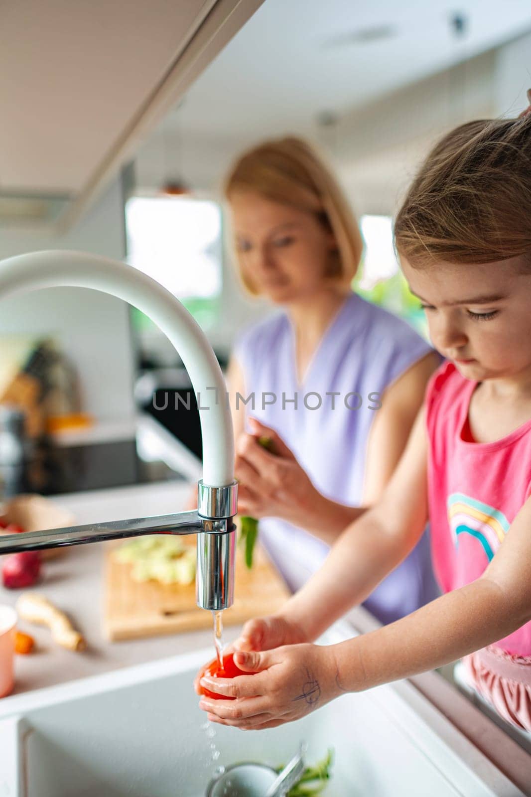 Young child washes vegetables while adult prepares food.