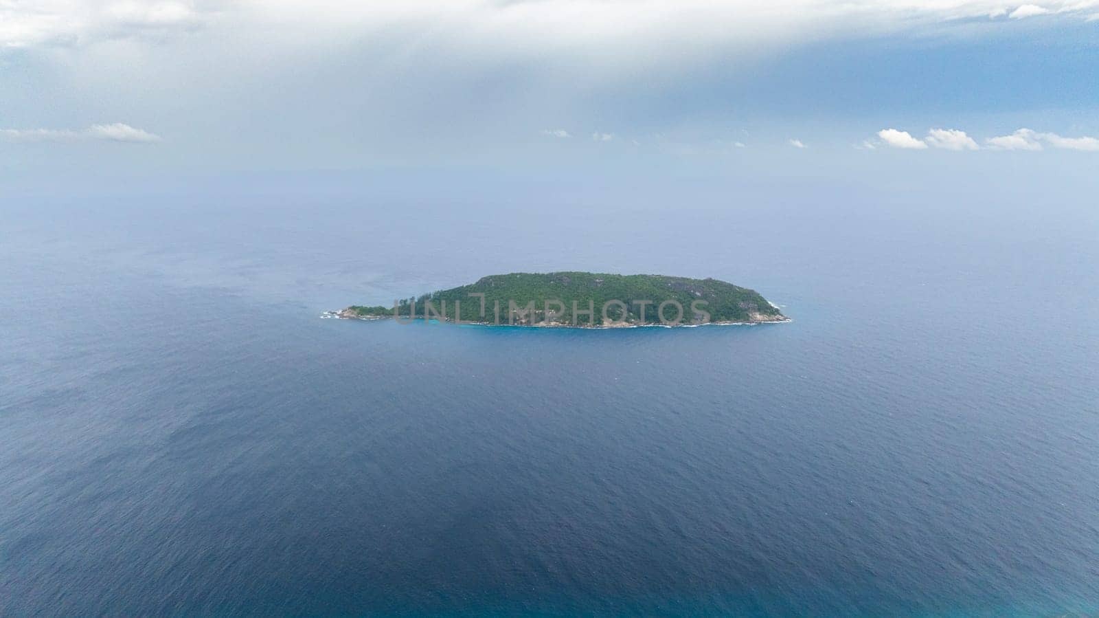 Green island surrounded by vast expanse of blue sea under overcast sky. Seychelles, Conception Island.