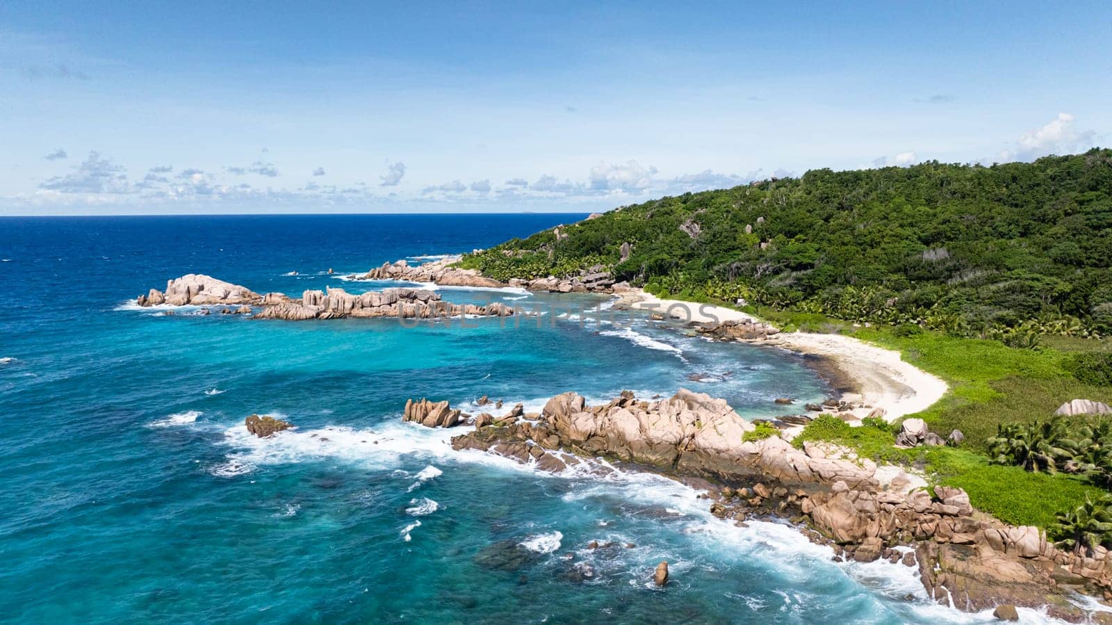 Tropical greenery meets clear turquoise ocean waters, with rocky outcrops along the coastline. La Digue, Seychelles.