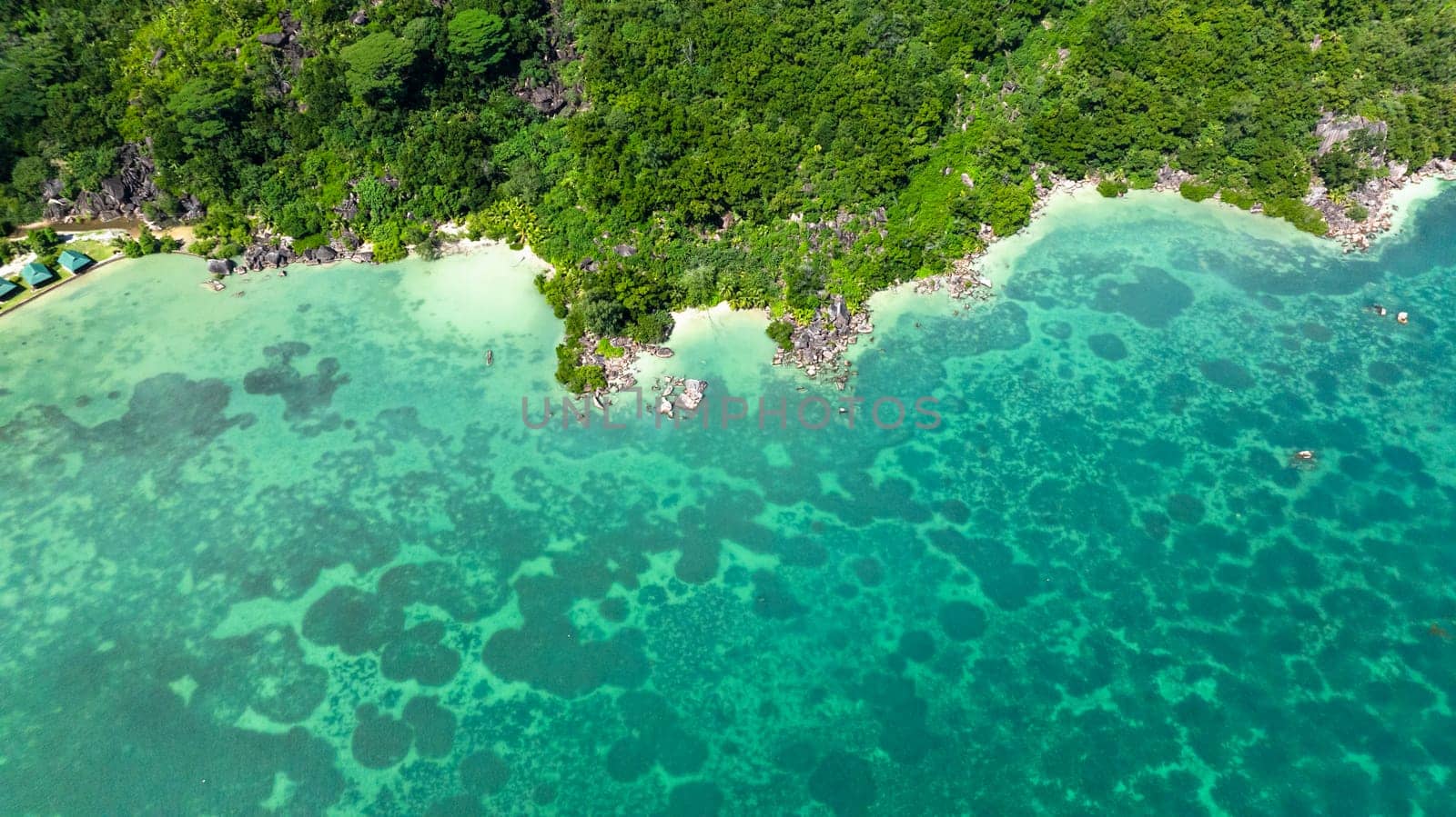 Aerial view of clear turquoise waters gently meeting a lush green coastline with rocky outcrops. Praslin, Seychelles.
