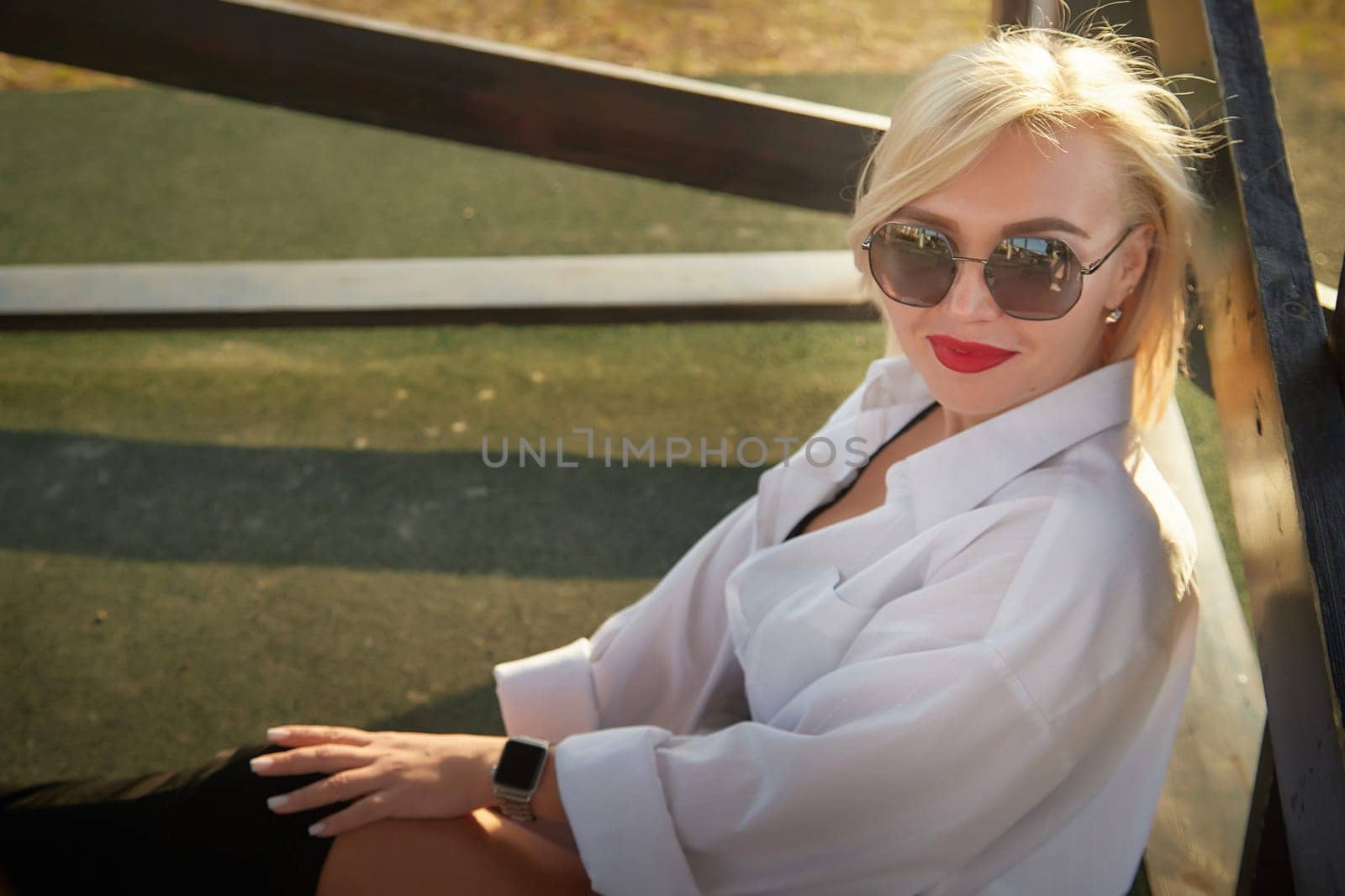 A woman poses comfortably outdoors wearing a stylish outfit and sunglasses, enjoying the warm sunlight in the late afternoon.