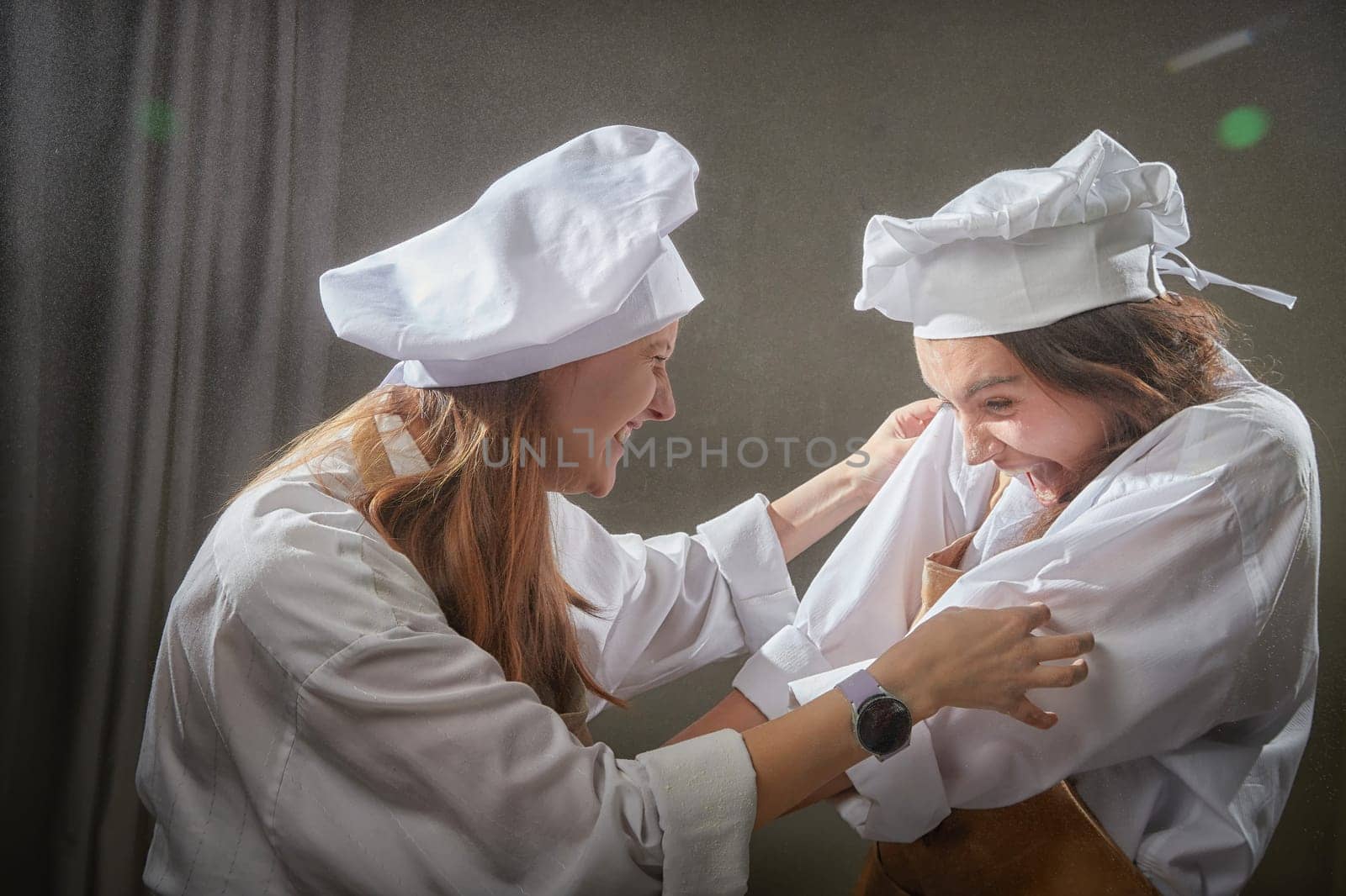 Two female friends laugh and playfully toss flour while dressed as chefs, enjoying a lively moment during their fun cooking photoshoot indoors.