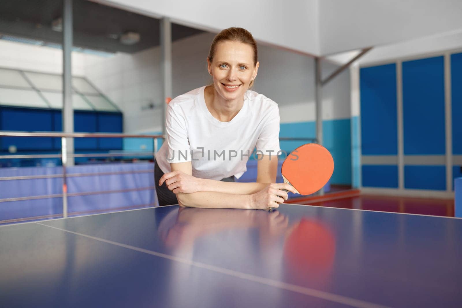 Smiling young woman holding racket in hand leaning over tennis table waiting for game start