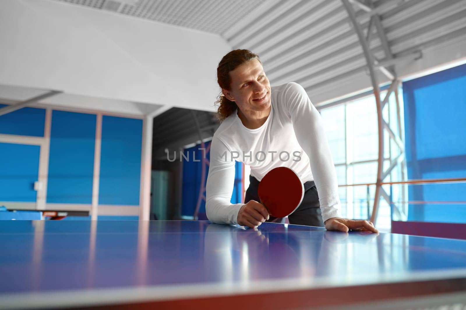 Satisfied sportsman having rest after training sitting on tennis table. Portrait of young man holding racket in hand