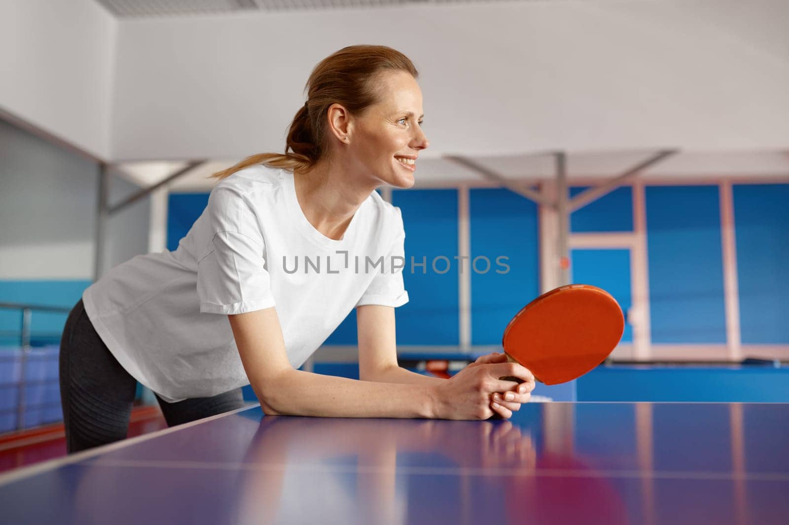 Happy smiling young woman holding racket in hand leaning over tennis table waiting for game start
