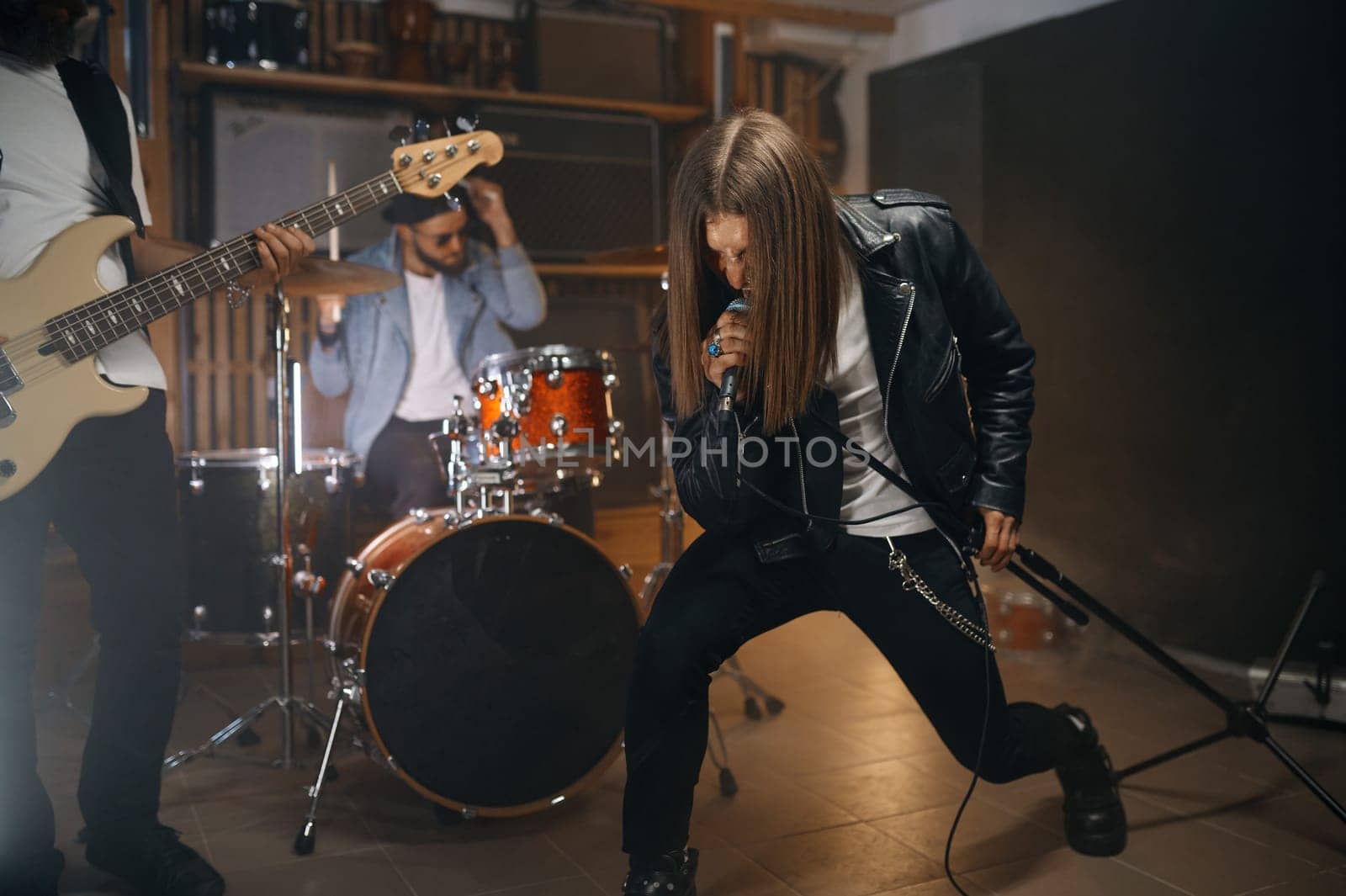 Expressive rocker singing song in microphone while his band playing heavy metal on drum and electric bass guitar. Young guys at rehearsal repetition in home studio
