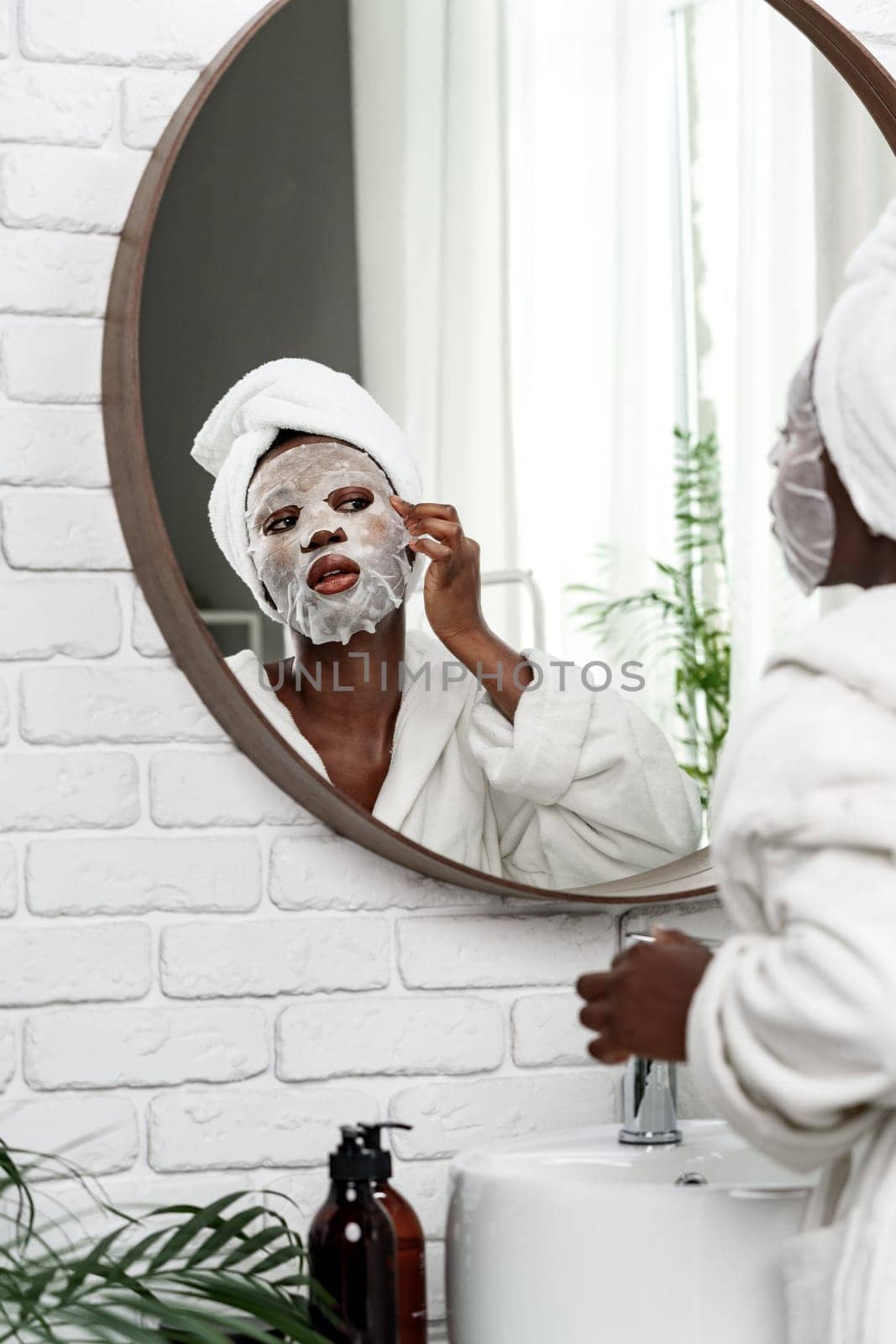 A person applies a facial mask while looking at their reflection in a round mirror. The bright bathroom features plants and a modern aesthetic, creating a serene atmosphere.