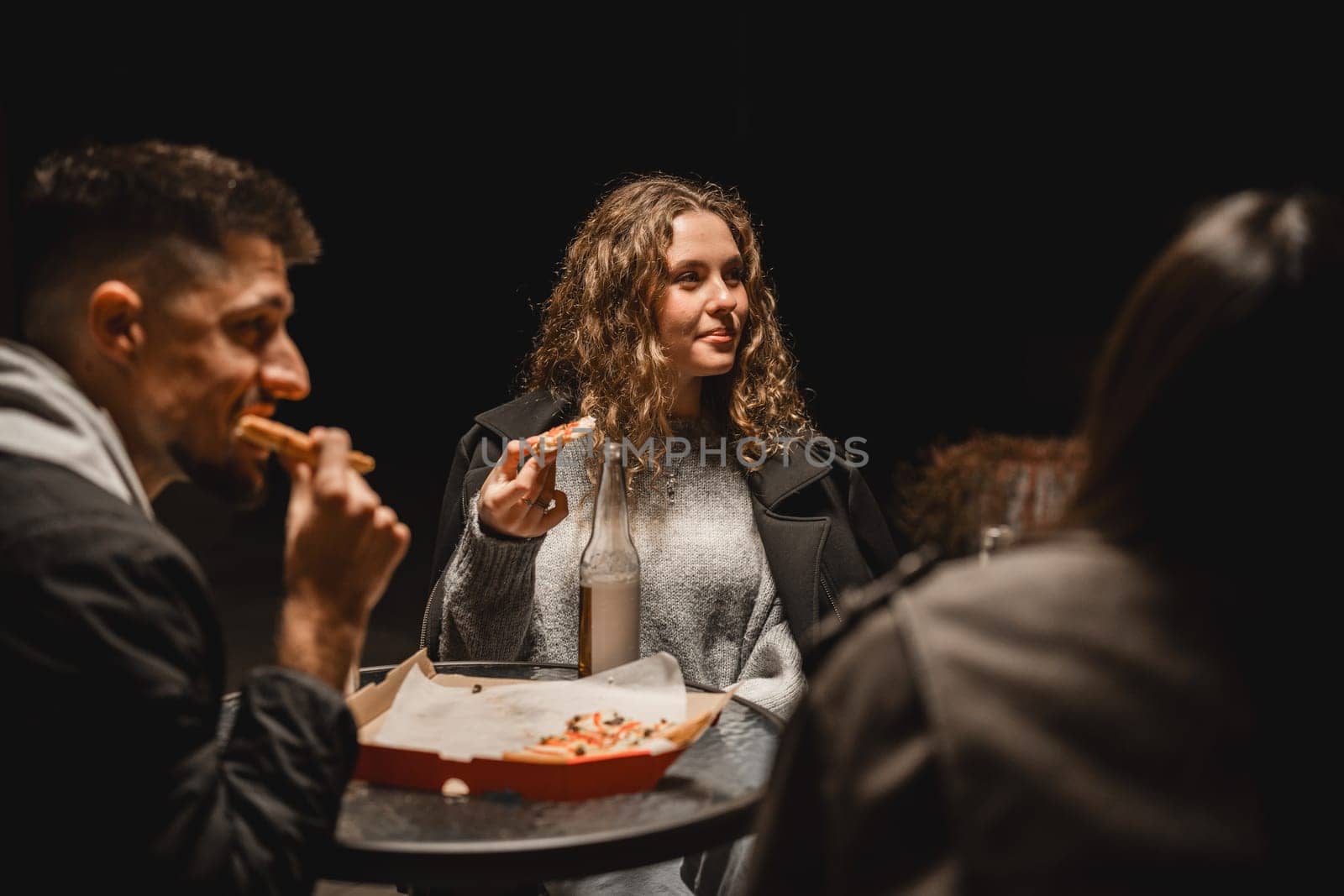 In a dimly lit setting, a group of friends shares a pizza, chatting and laughing. One person is holding a slice while others enjoy their drinks, creating a warm atmosphere.