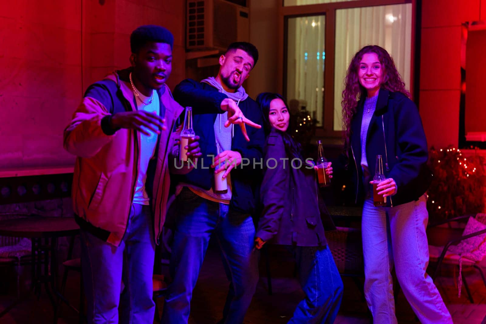Four friends enjoy a fun evening together in a cozy indoor space, illuminated by vibrant colorful lights while holding drinks and posing playfully.
