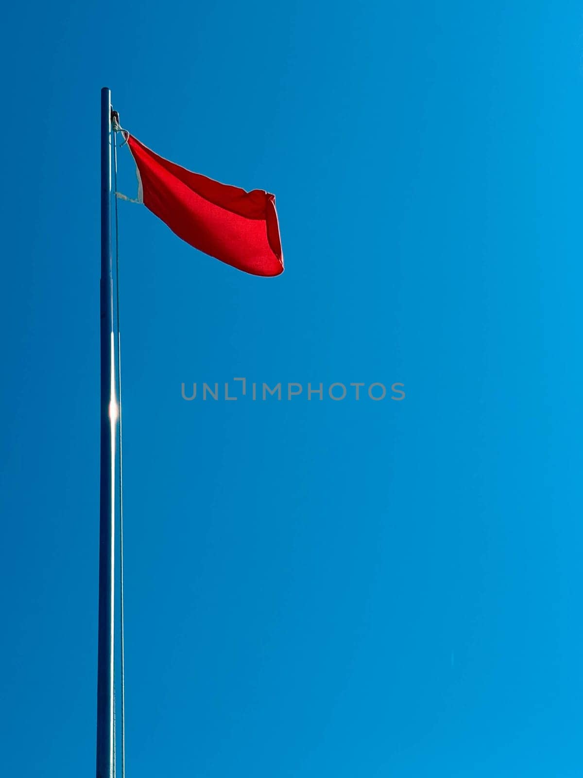 A red flag flutters in the wind in the blue sky by the sea during the day. Danger sign in the form of a red flag.