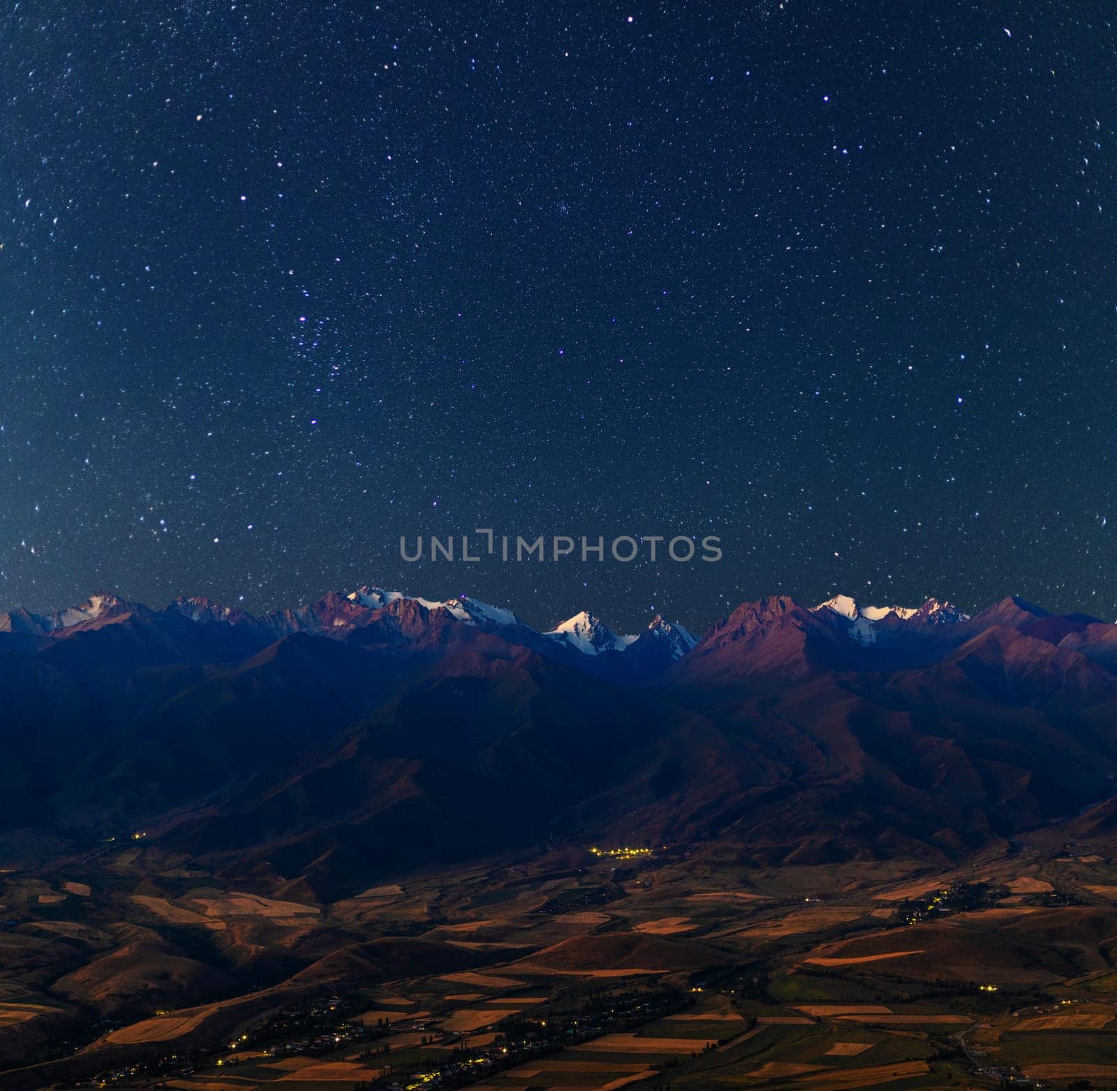 A breathtaking starry night sky illuminates the rugged, snow-capped mountains of Kyrgyzstan in summer, with a serene valley and scattered village lights below.