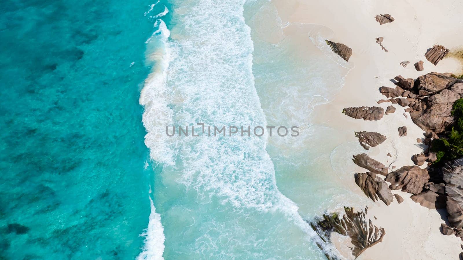 Turquoise ocean waves and white sands meeting rugged rock formations along the coast. La Digue, Seychelles.