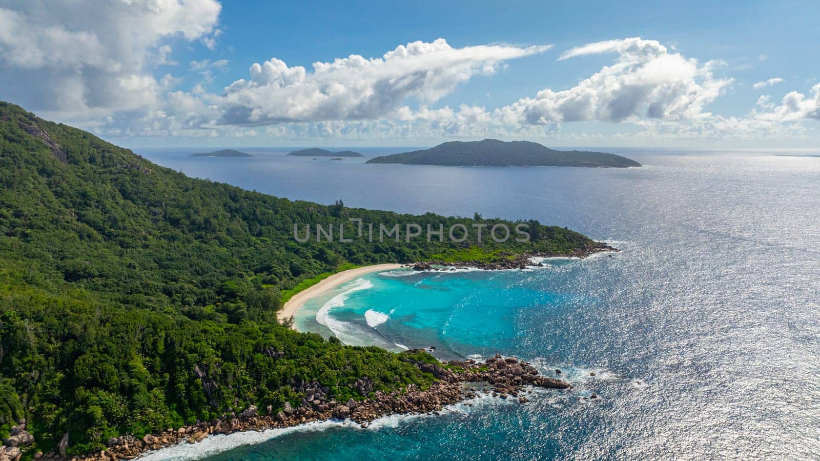 Waves gently roll onto a white sandy beach, bordered by lush green vegetation. La Digue, Seychelles.