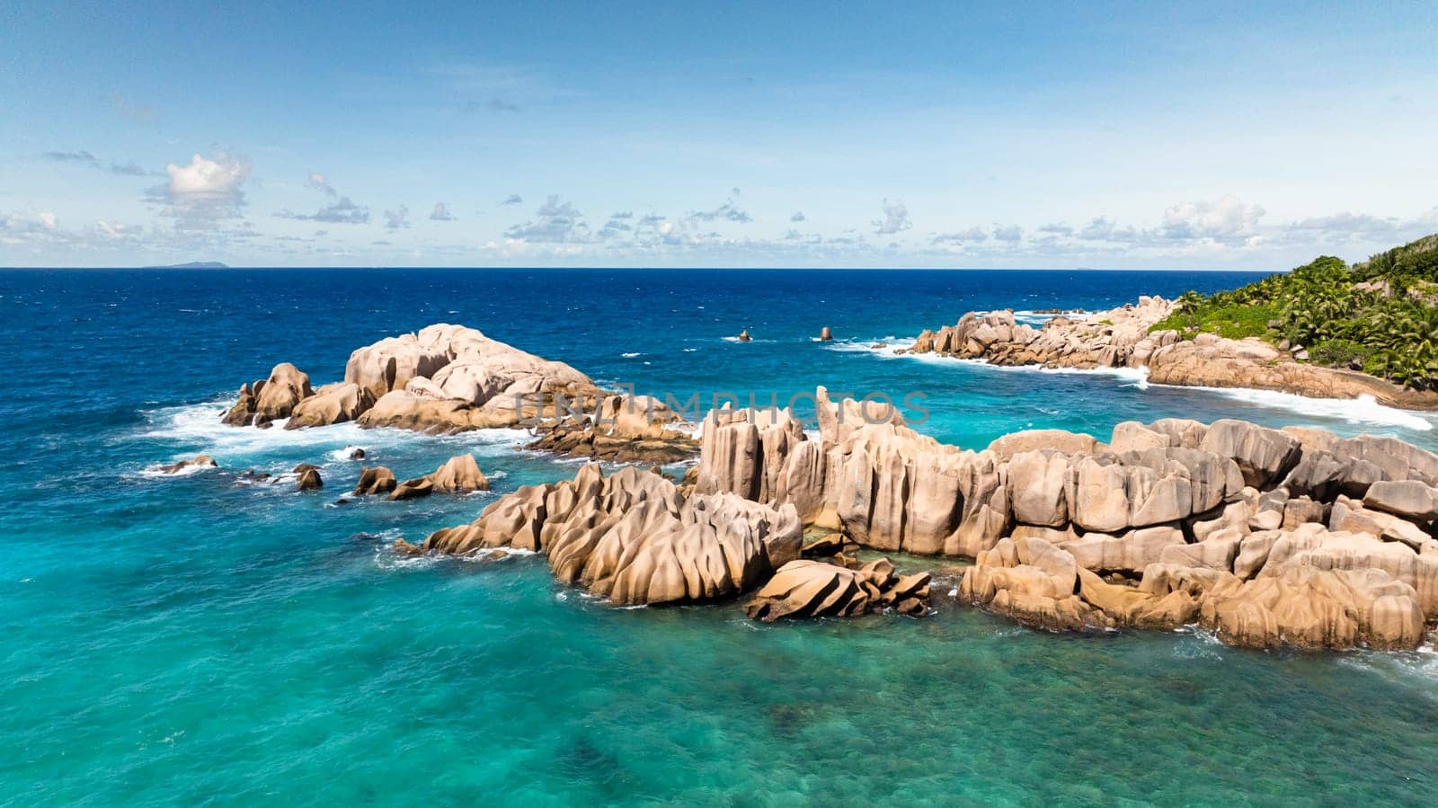Rocky coastline under a bright blue sky with some clouds in the distance. La Digue, Seychelles.